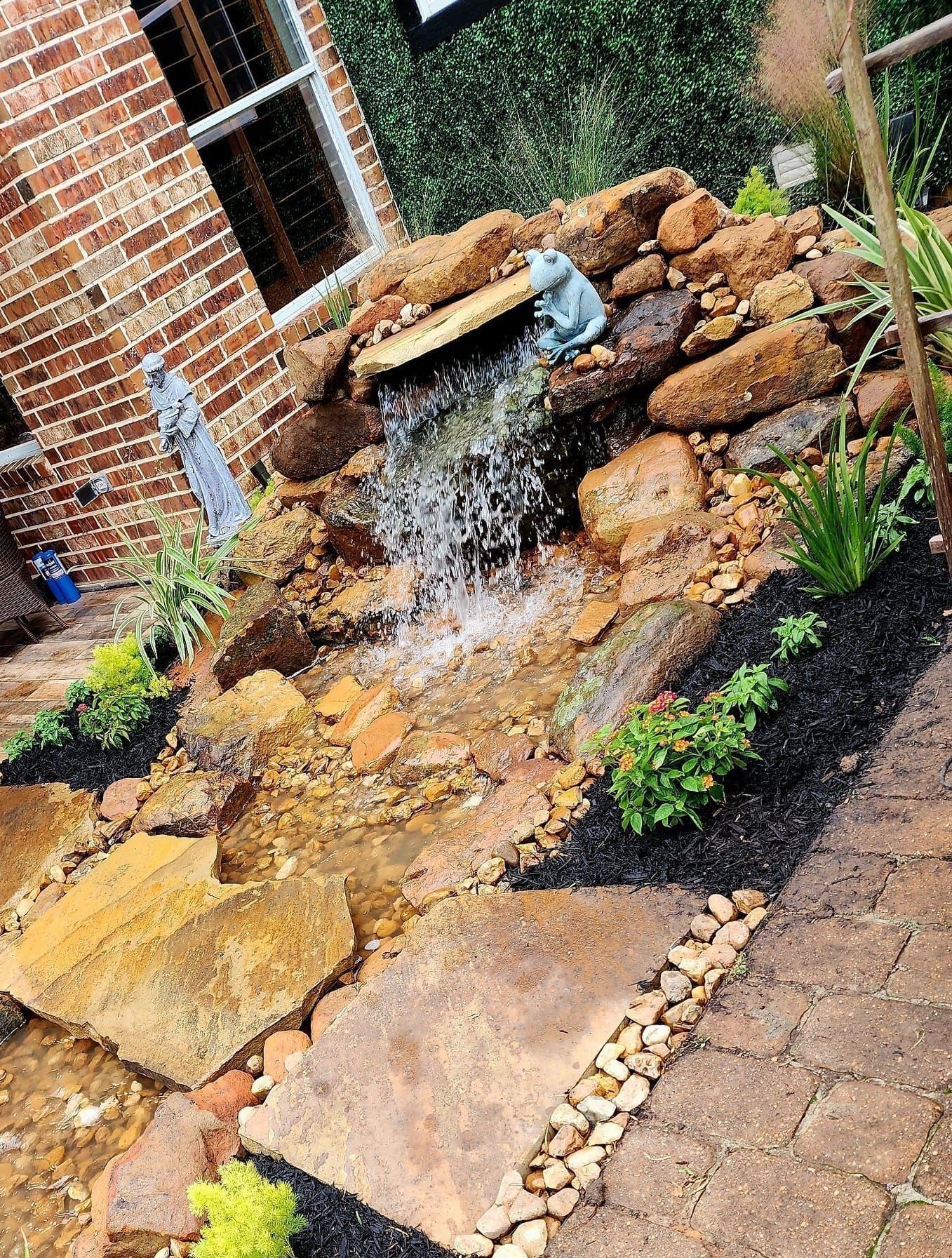 Small backyard waterfall feature with rocks and greenery, next to brick building.