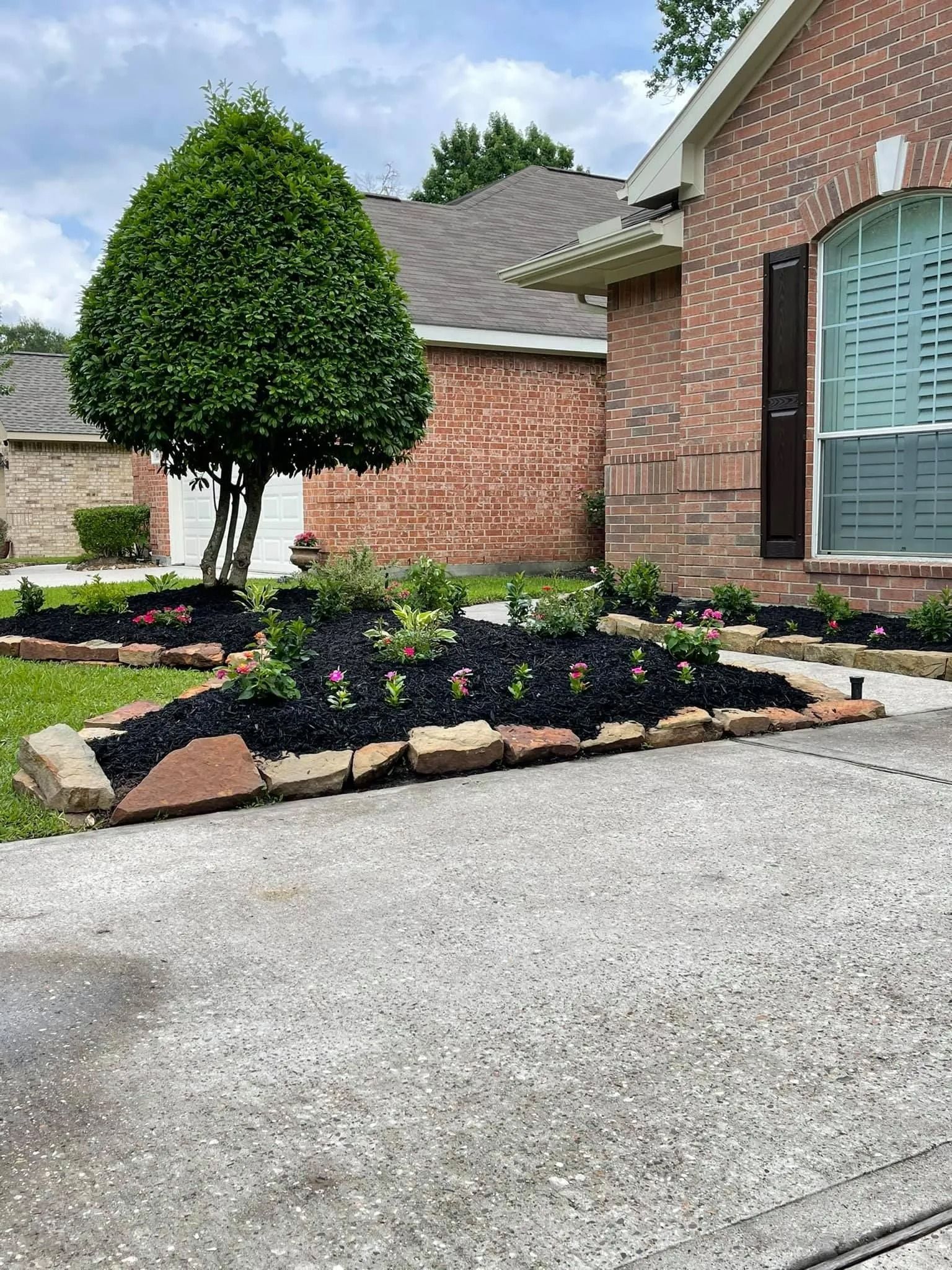 A landscaped yard with a brick house. Mulched flower beds bordered by stone, green tree, and driveway.
