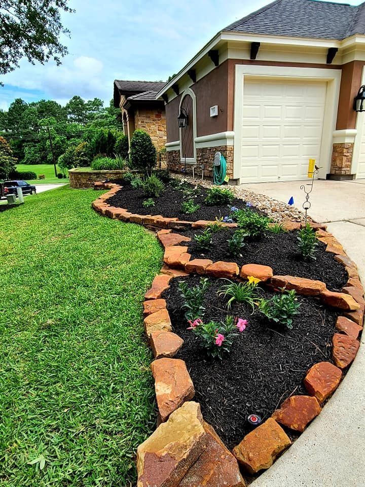 A well-manicured front yard with a mulch flower bed bordered by reddish-brown stones, alongside a driveway and beige house.