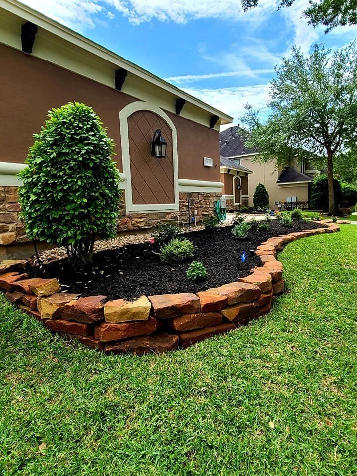A house with a brick-edged flower bed filled with black mulch, green plants, and a small, round tree.