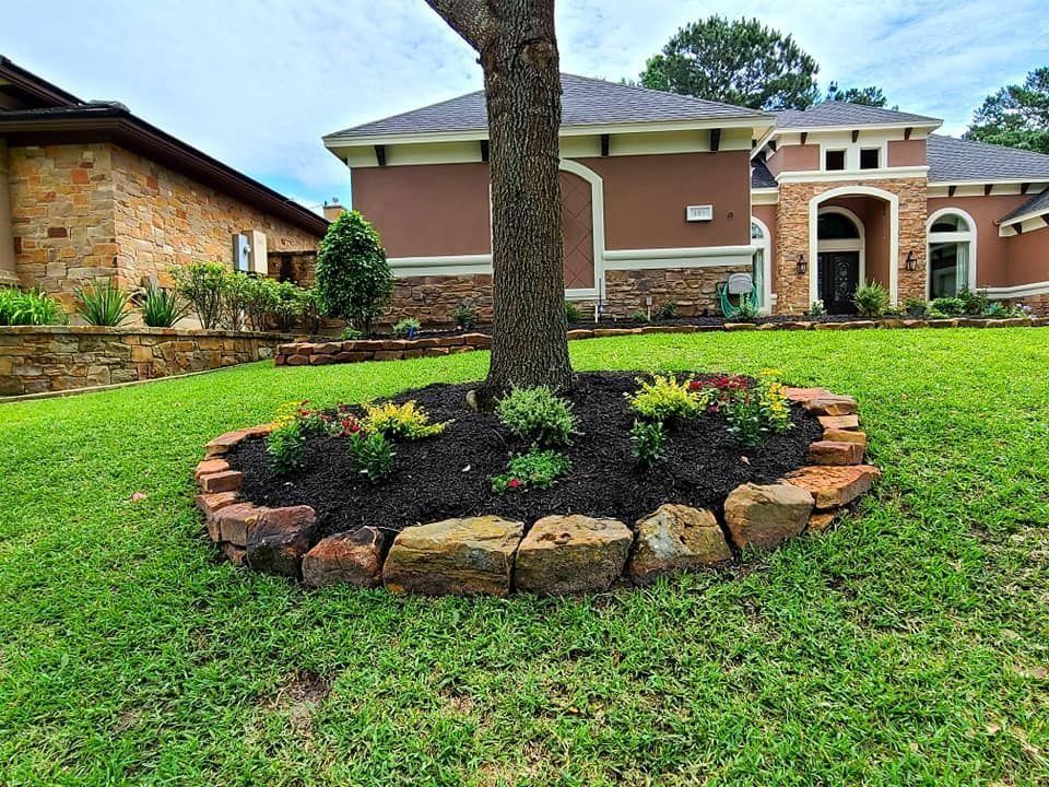 Tree with flower bed, surrounded by stone border, in front of a house on a green lawn.