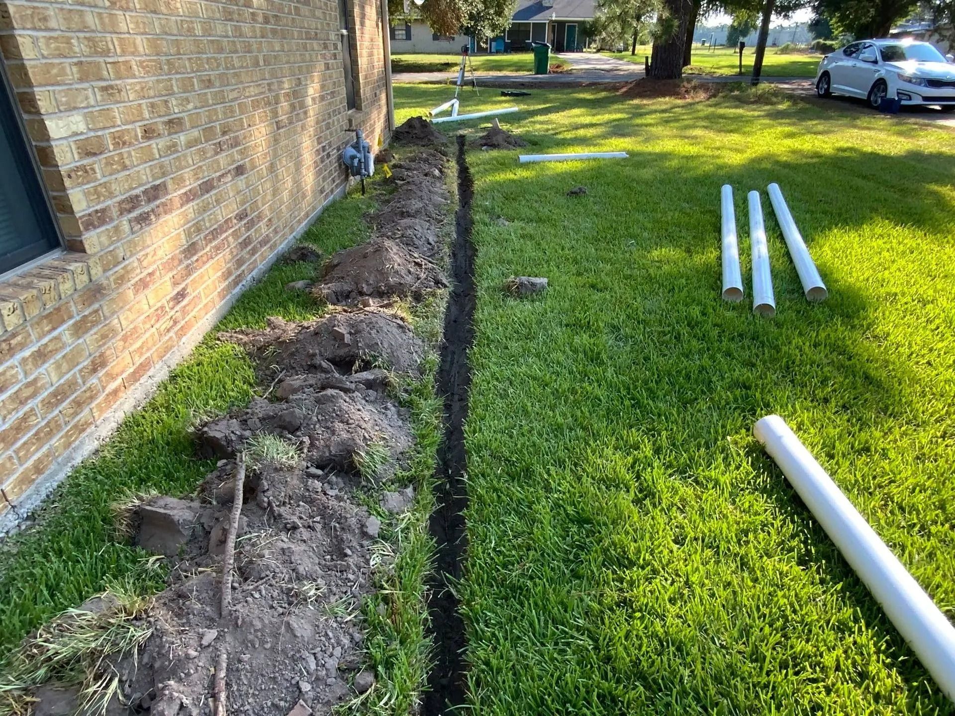 A trench dug in green grass along a brick building; white pipes lie nearby.