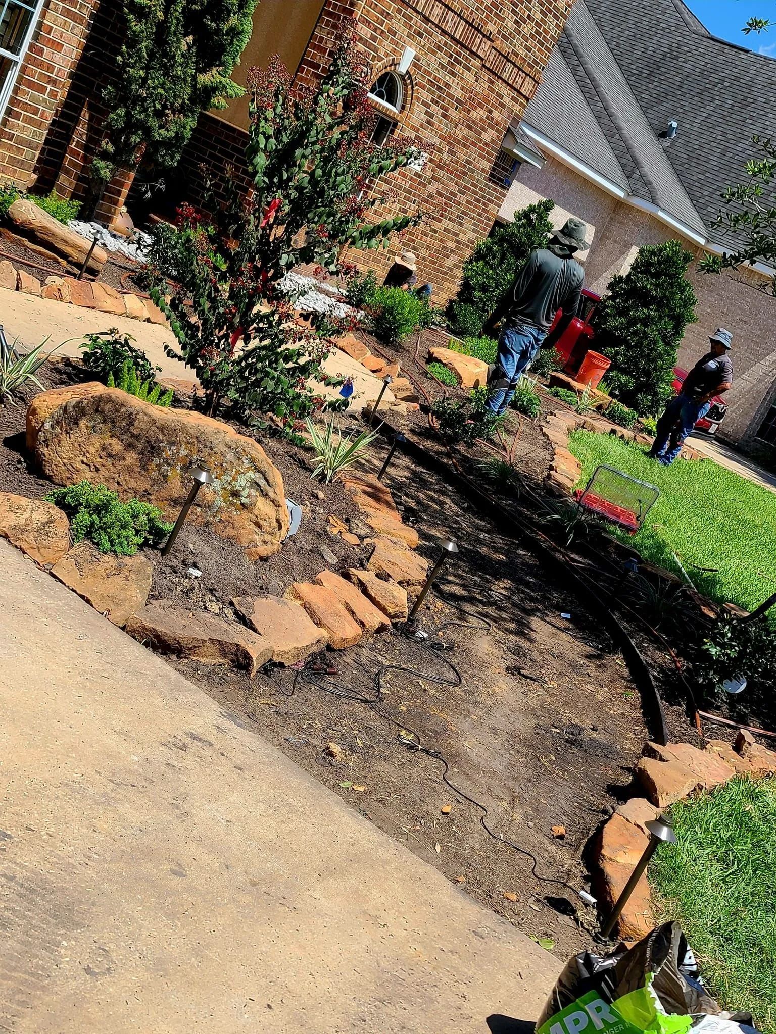 Workers landscaping a yard with a brick-edged flower bed, beside a concrete walkway, next to a home.