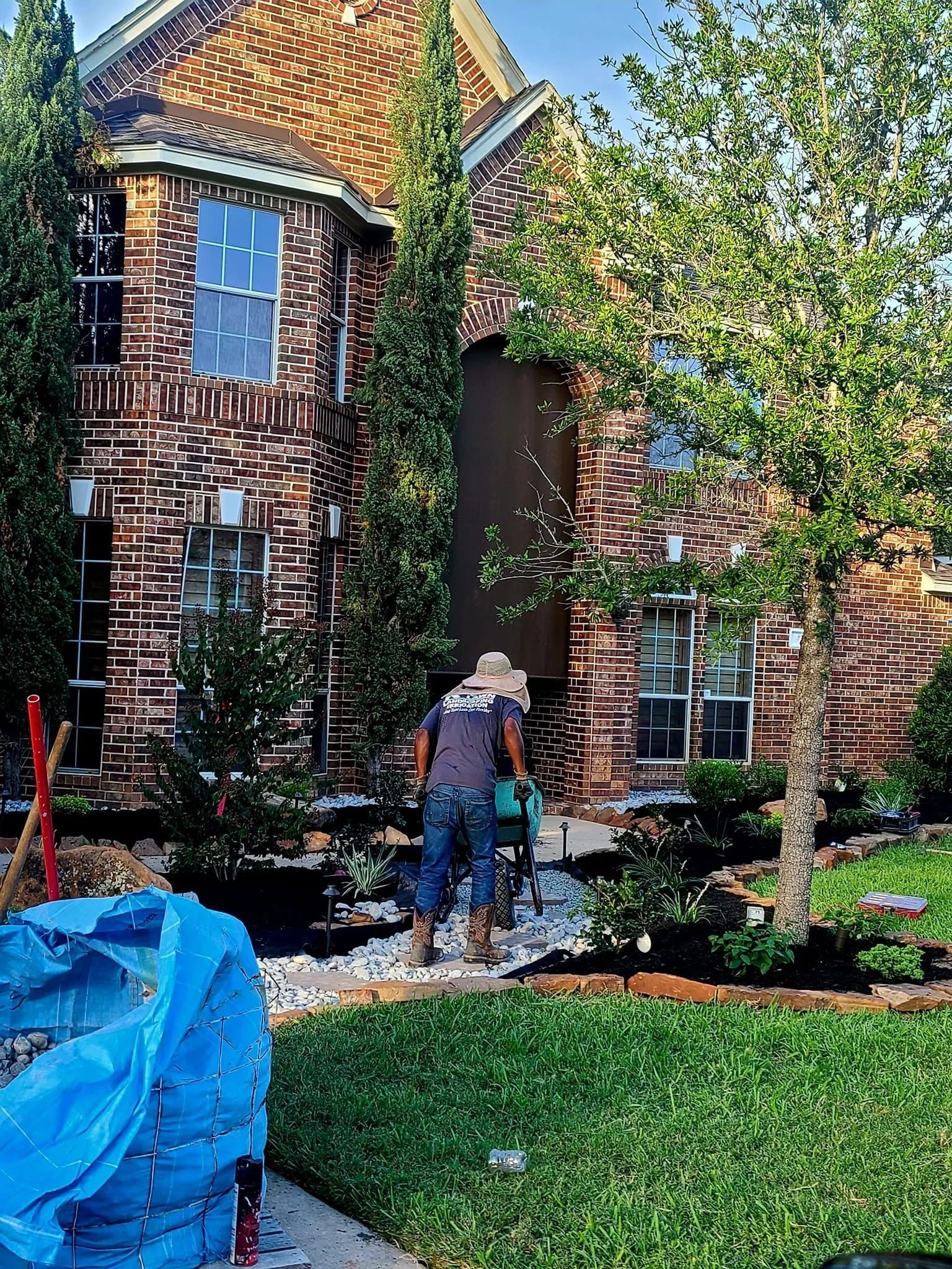Person using a tool in a landscaped yard in front of a brick house with large windows; blue tarp in the foreground.