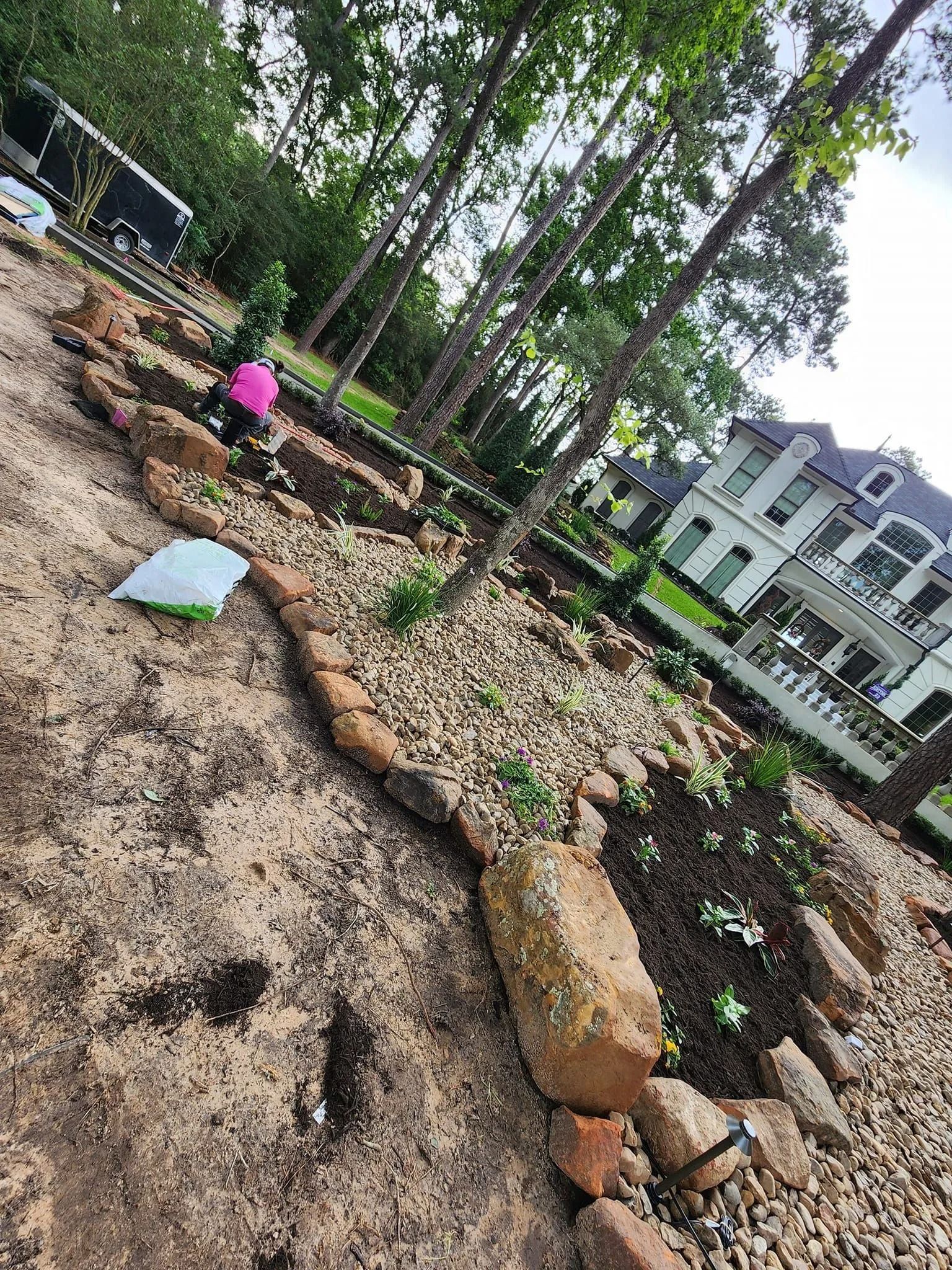 Person gardening in a rock and brick bordered bed; trees and large house in background.