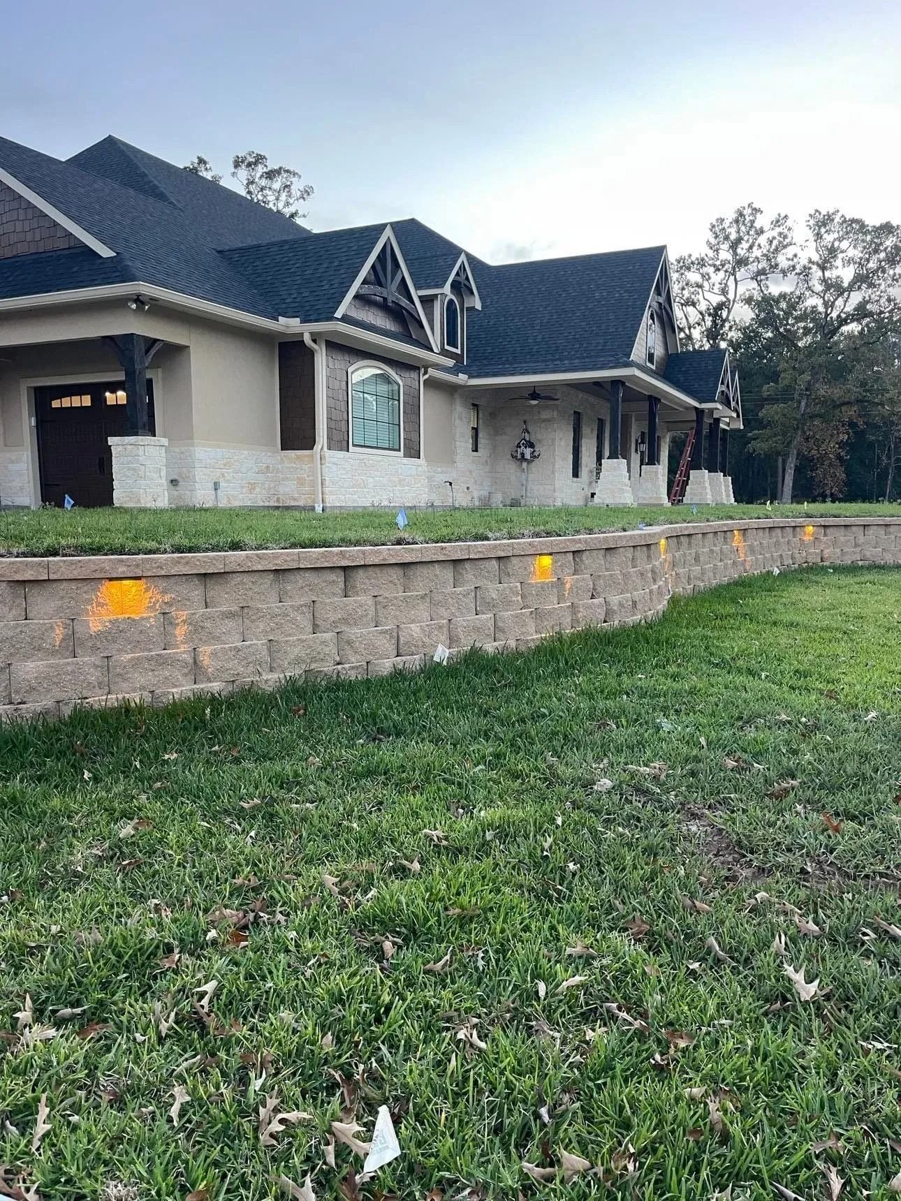 A large, beige-colored house with a black roof sits behind a retaining wall with built-in lights.