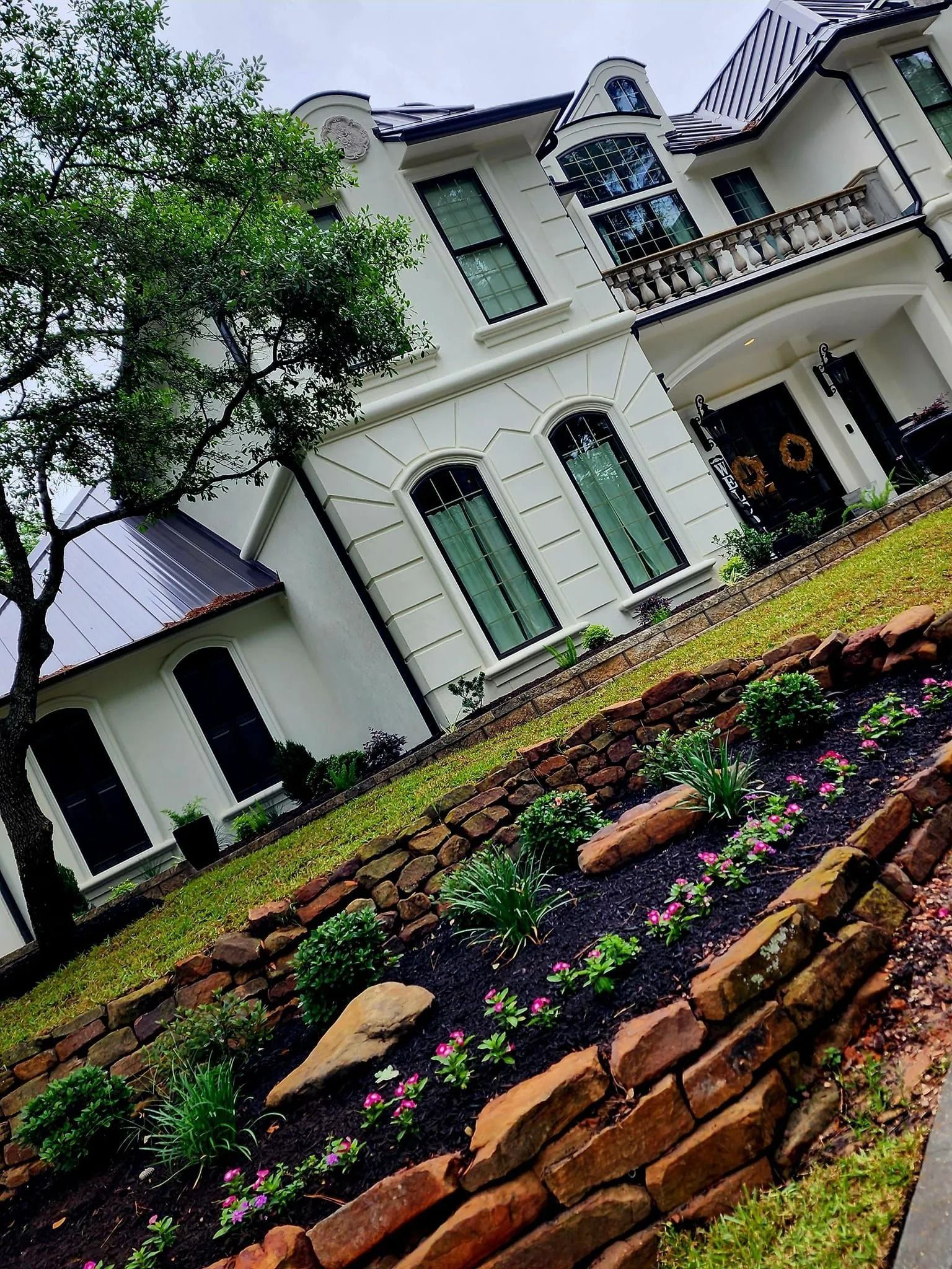 White mansion with brick flower bed, lush green lawn, and dark roof.