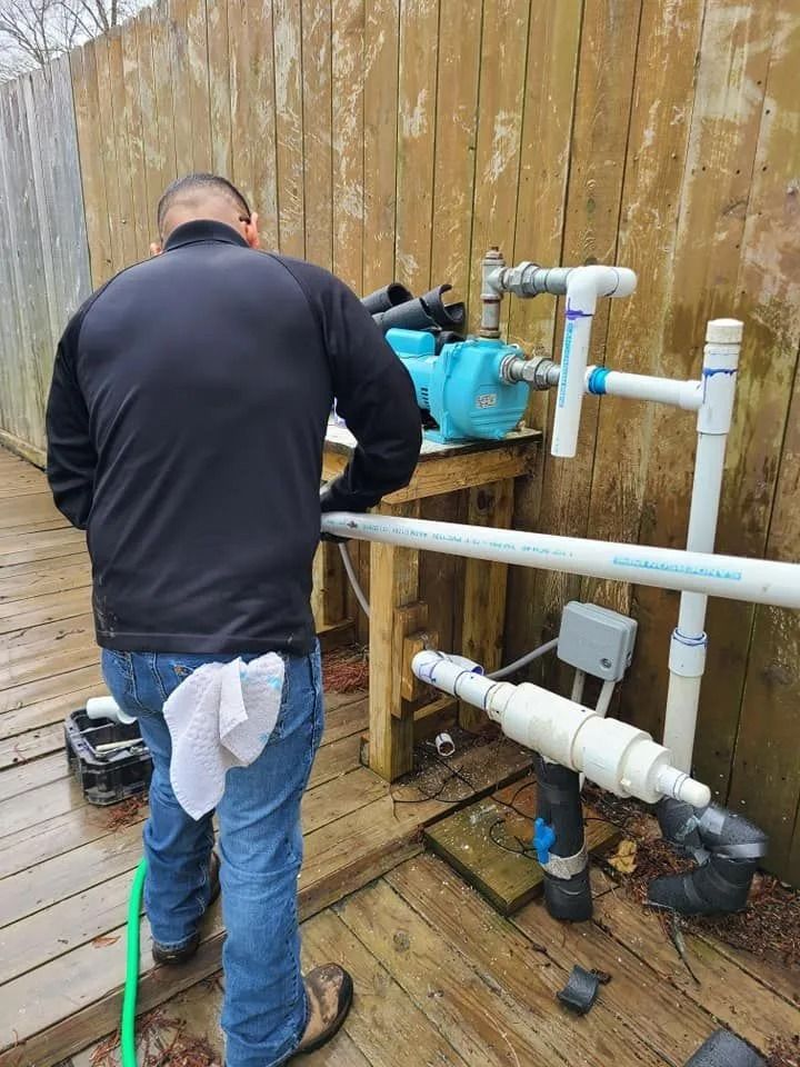 Person working on plumbing, next to a blue pump and white PVC pipes on a wooden deck.