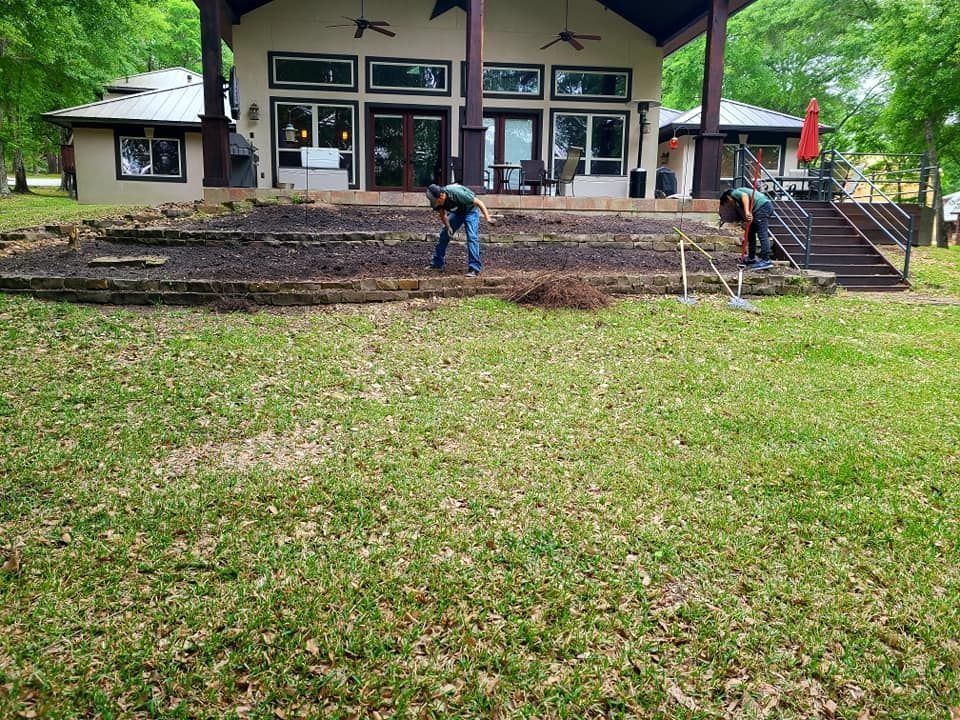 Two people working on a tiered garden in front of a house. One is digging, the other raking. Green lawn.