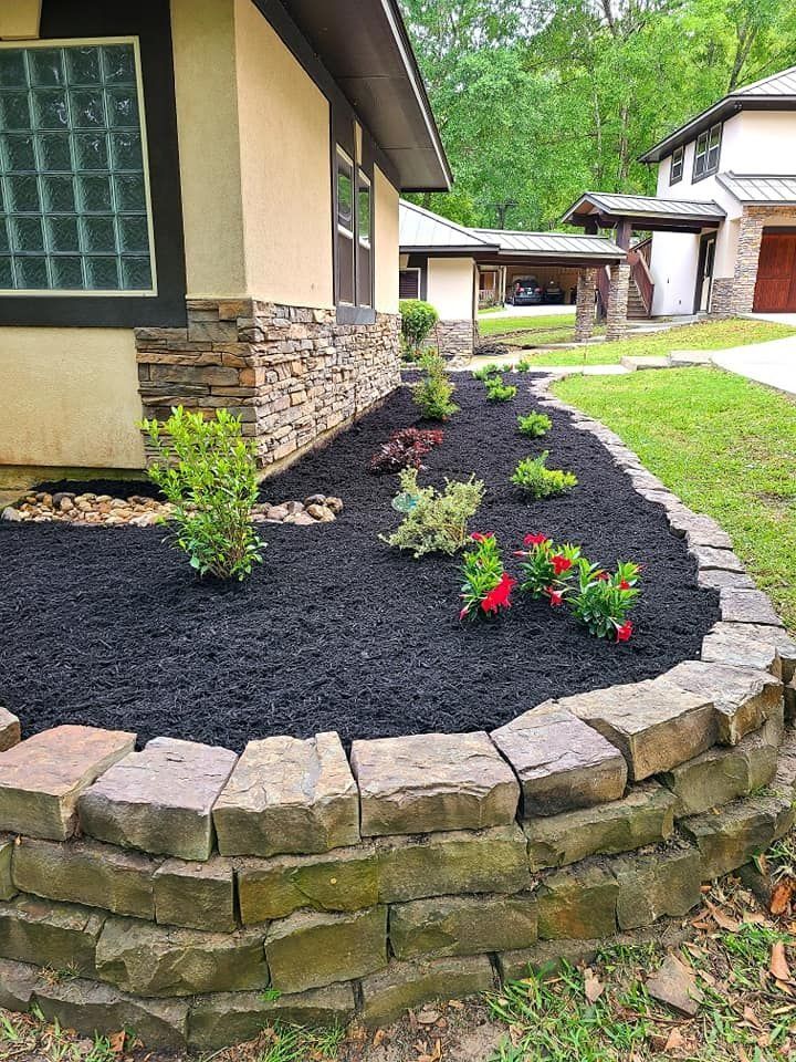 Stone-edged flower bed with black mulch and green plants in front of a house.