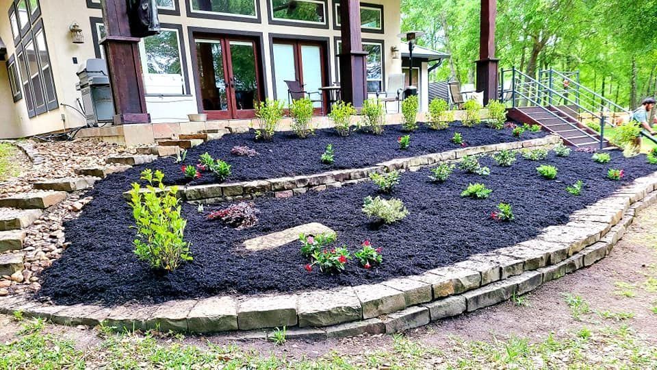 Landscaped tiered garden bed with black mulch, various plants, and stone retaining walls.