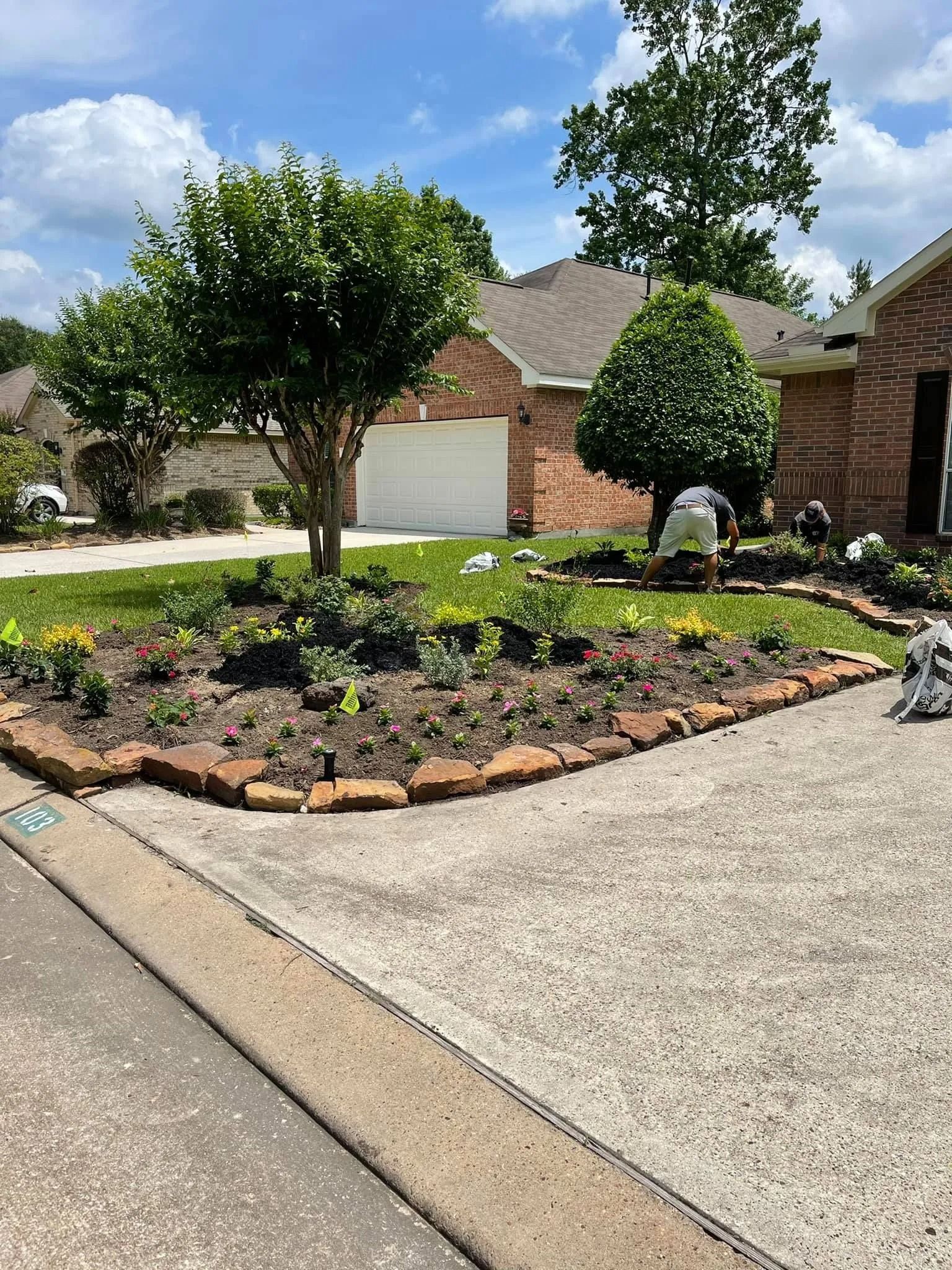 A house with a brick facade and a landscaped garden bed. A person works in the garden, and the sky is sunny.