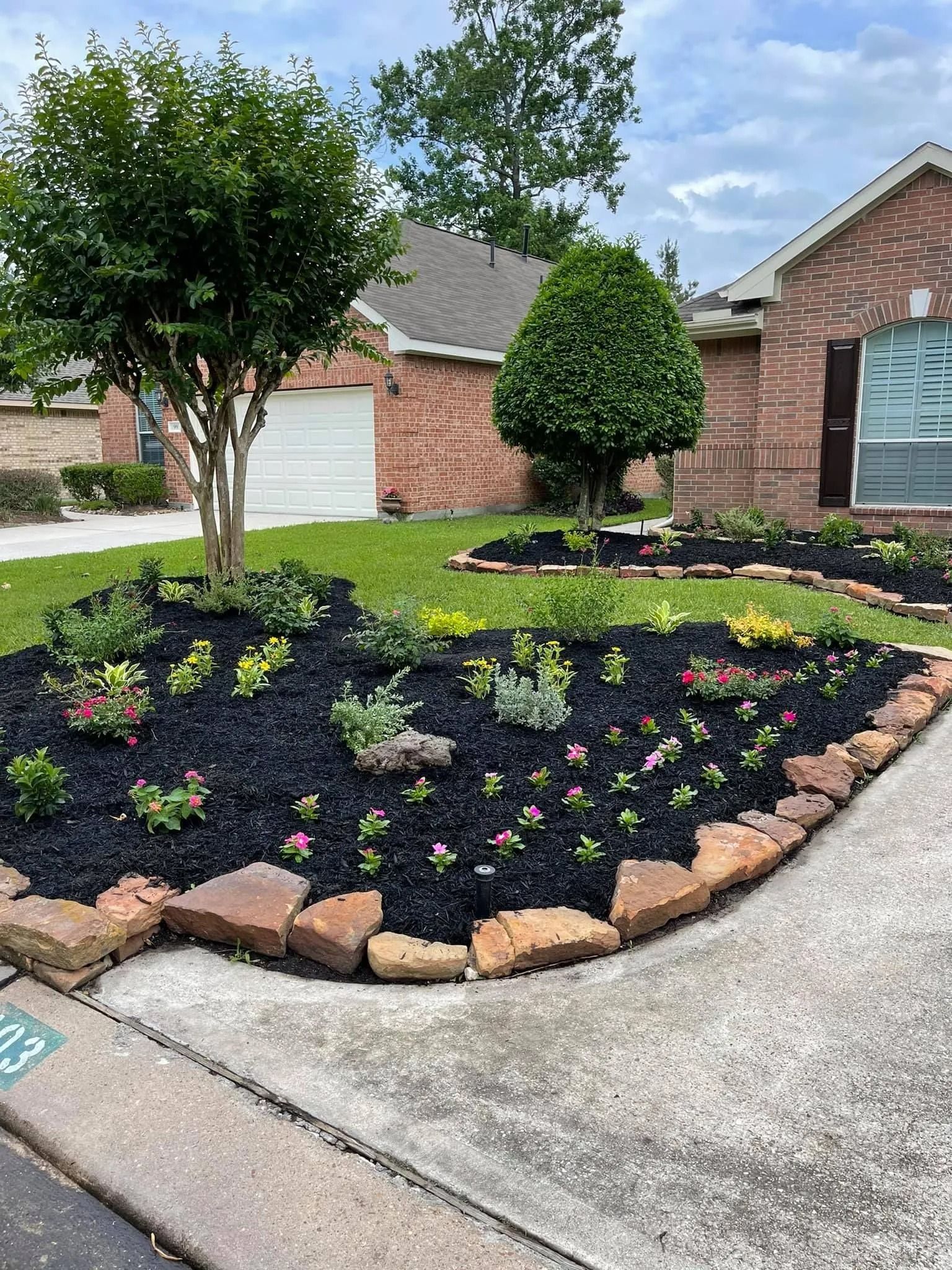 Well-maintained front yard with flower beds, mulch, brick edging, and trees in front of a brick house.