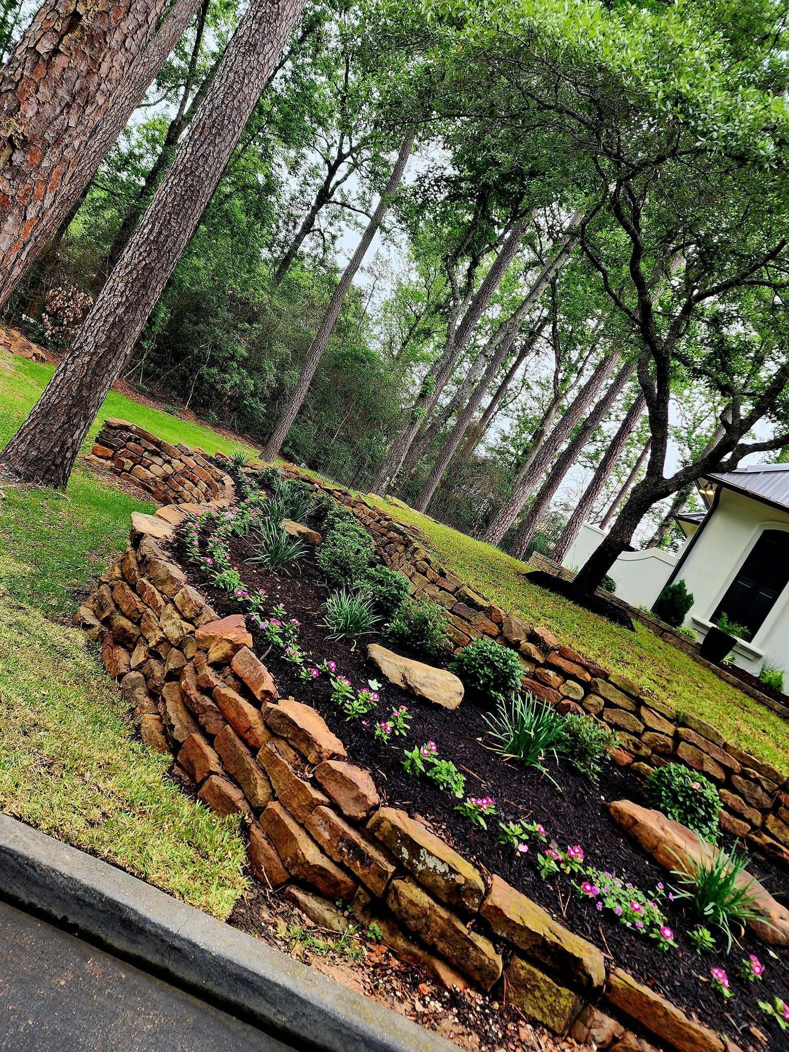 Stone-lined garden bed on a slope with green plants and small pink flowers, near trees and a white house.