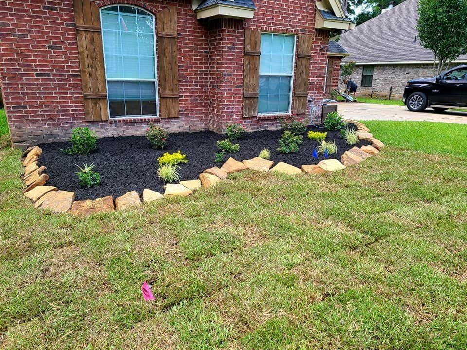 Flower bed in front of a brick house, bordered by stone.  Fresh mulch and plants. Green grass in foreground.