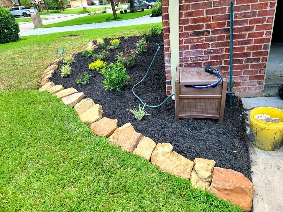 Flower bed with dark mulch, stone edging, near a brick wall, hose, and grass.