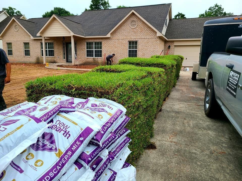 A house with a trimmed hedge, bags of soil, and a truck parked on the driveway; a person works in the yard.