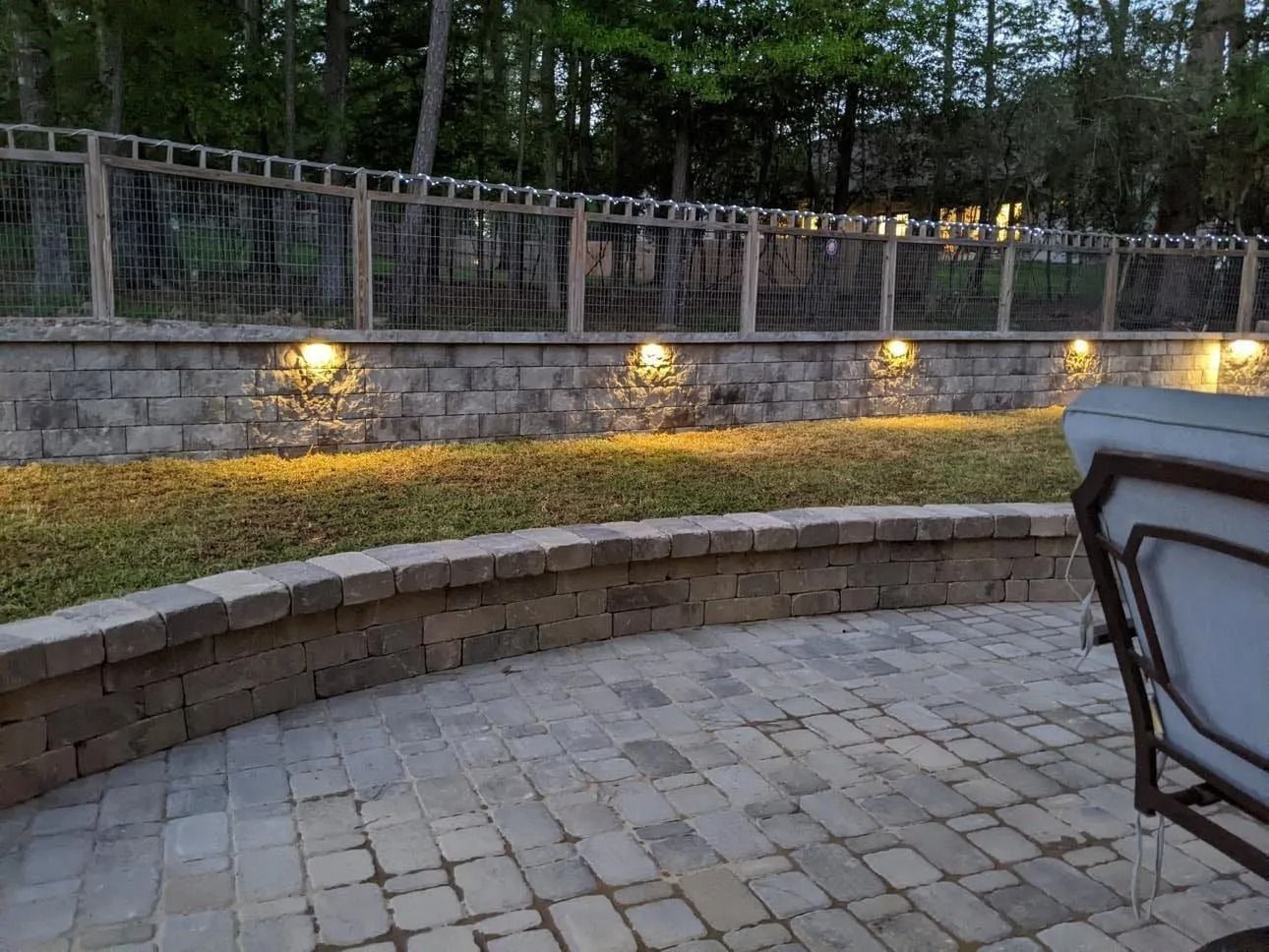 Stone patio with curved retaining wall, lawn, and lit stone wall with fence and trees in background.
