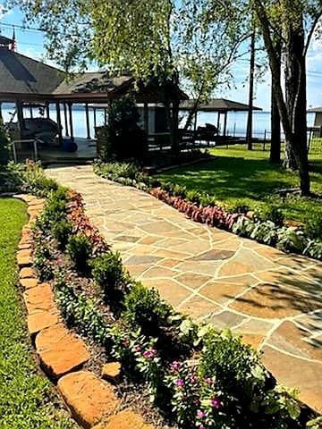 Stone path lined with flowers and shrubs leads to a lakefront house and gazebo on a sunny day.