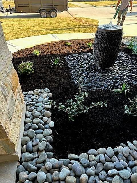 Front yard landscaping with a dark rock water fountain, mulch, and river rock borders.