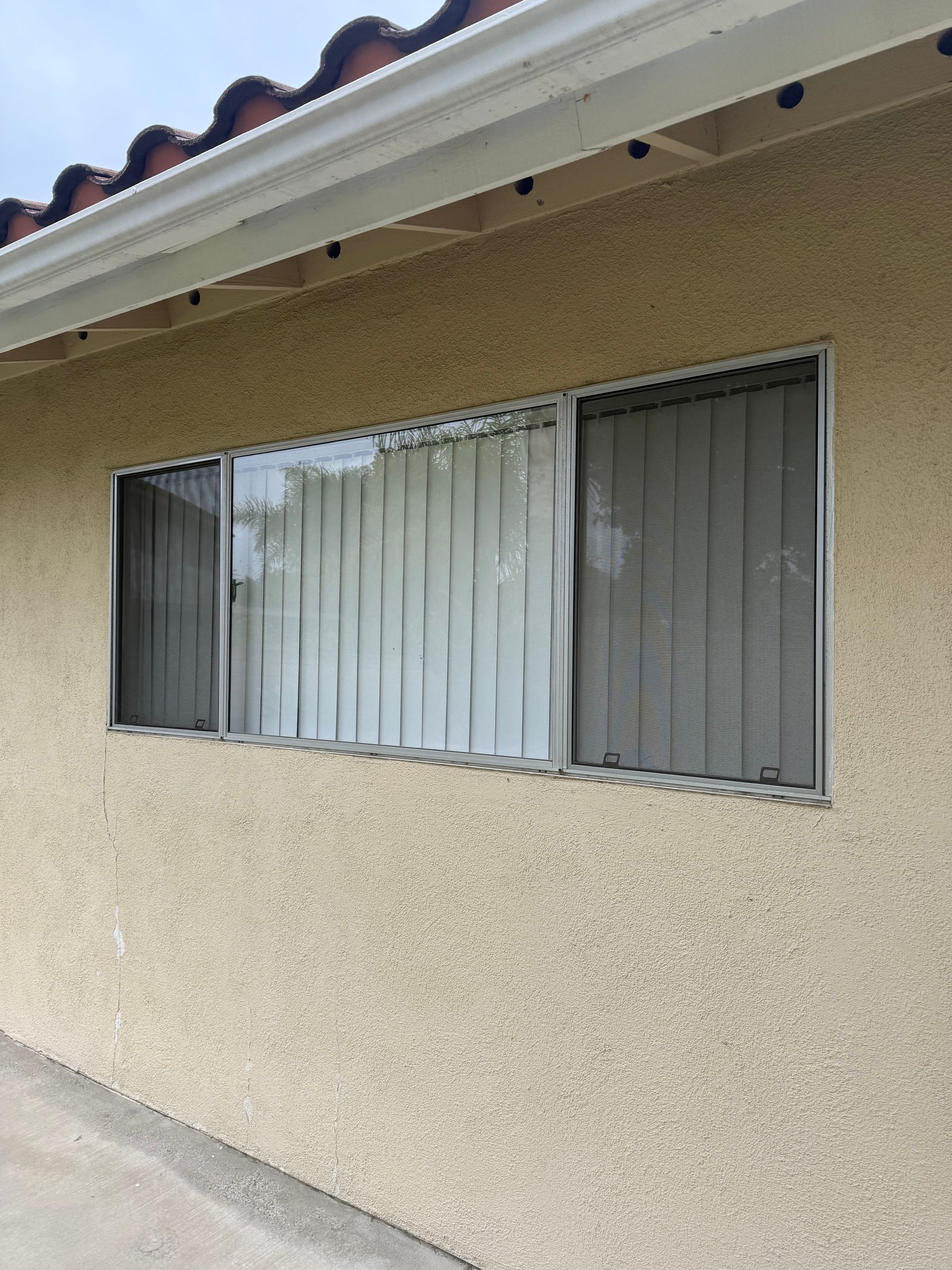 Exterior wall of a house with a window, white curtains, and closed gray blinds under a tiled roof.