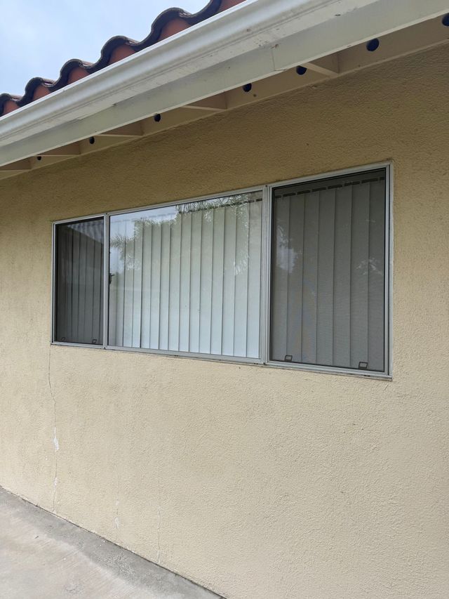Exterior wall of a house with a window, white curtains, and closed gray blinds under a tiled roof.