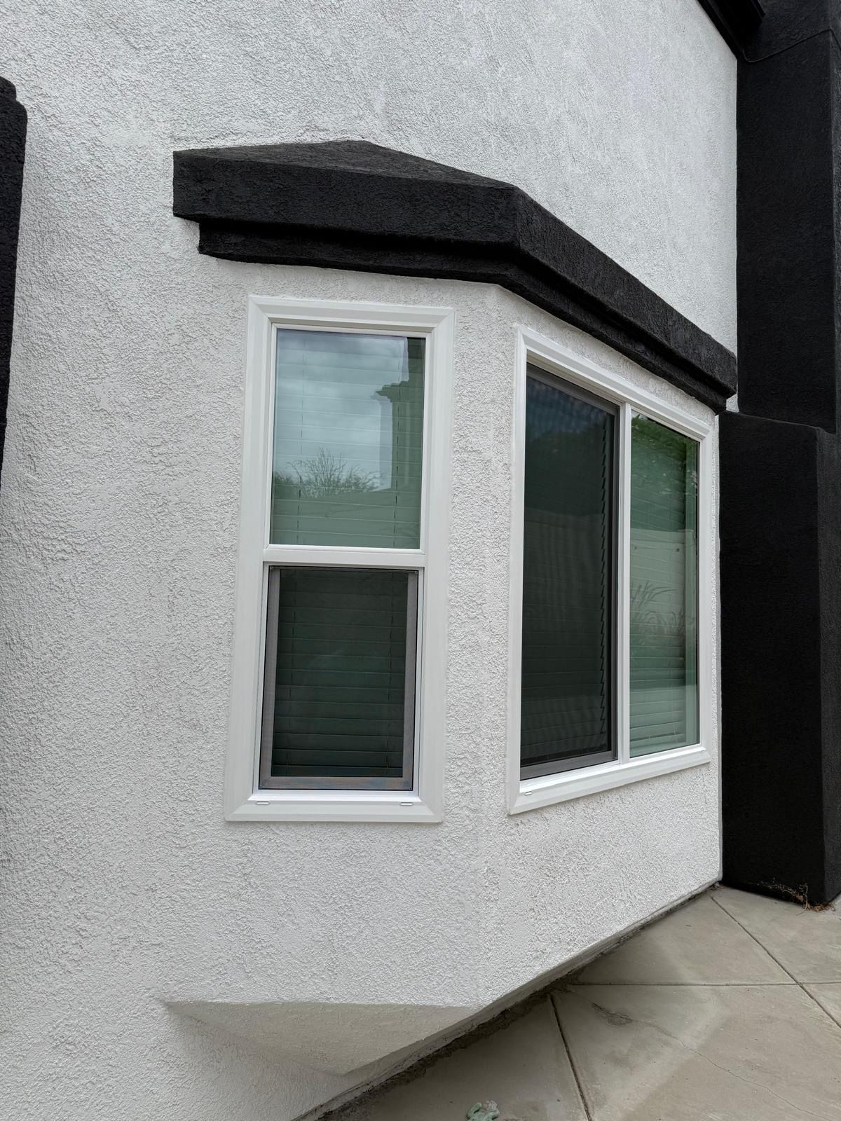 Small white stucco house wall with a bay window and black awning beside a paved walkway