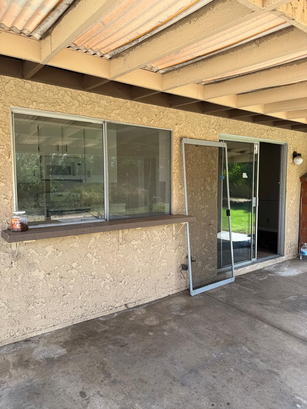 Covered patio with a large window, open screen door, and concrete floor next to a grassy yard