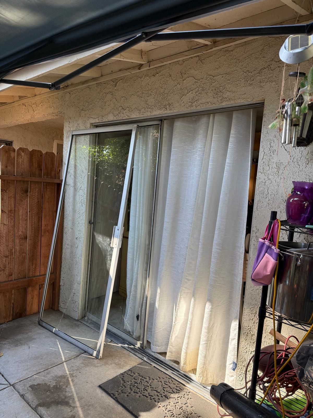 Patio sliding glass door with white curtains, leaning frame, and hanging planters beside a stucco wall