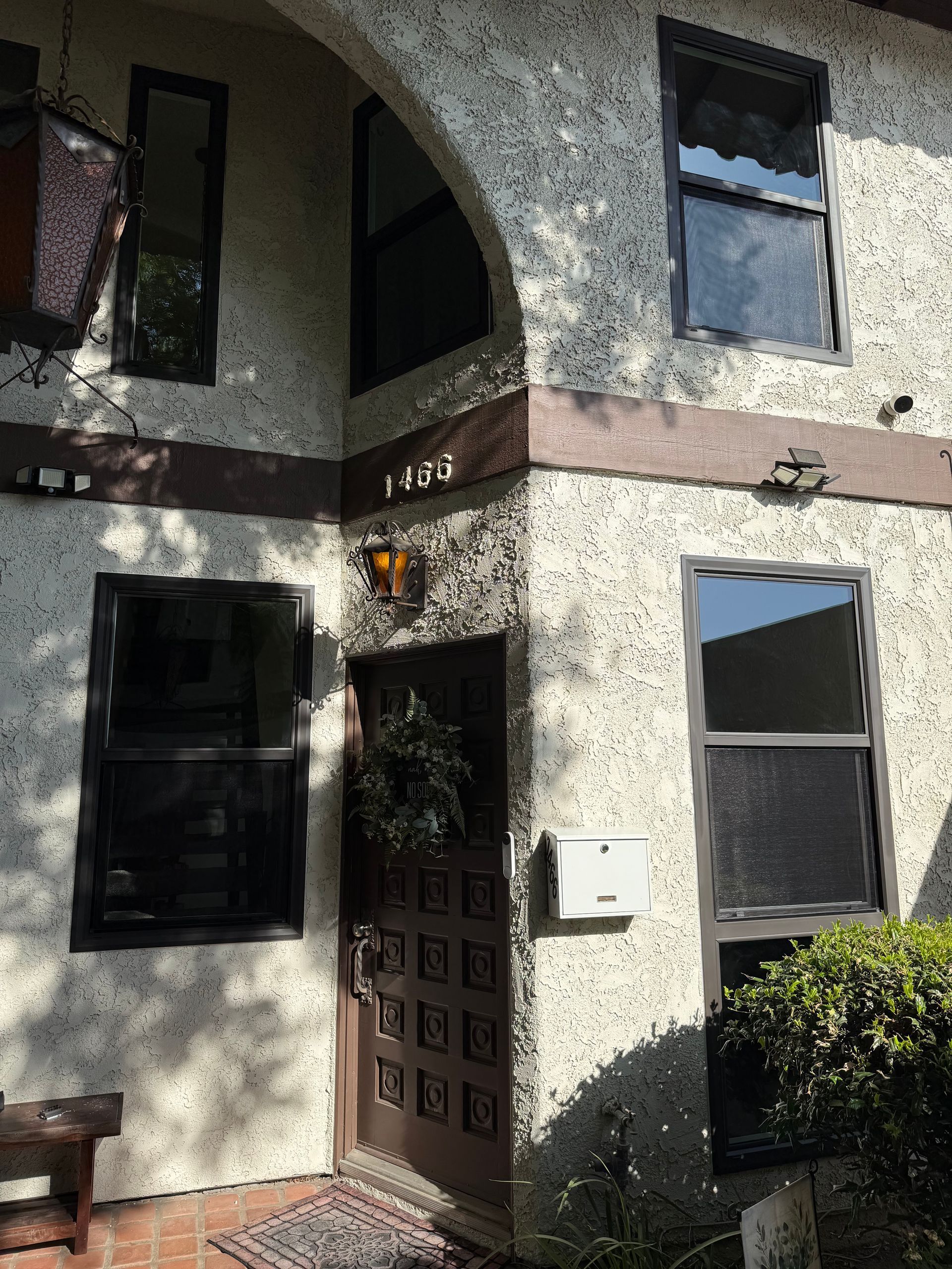 Two-story stucco house entrance with dark front door, windows, and a small shrub by the wall.