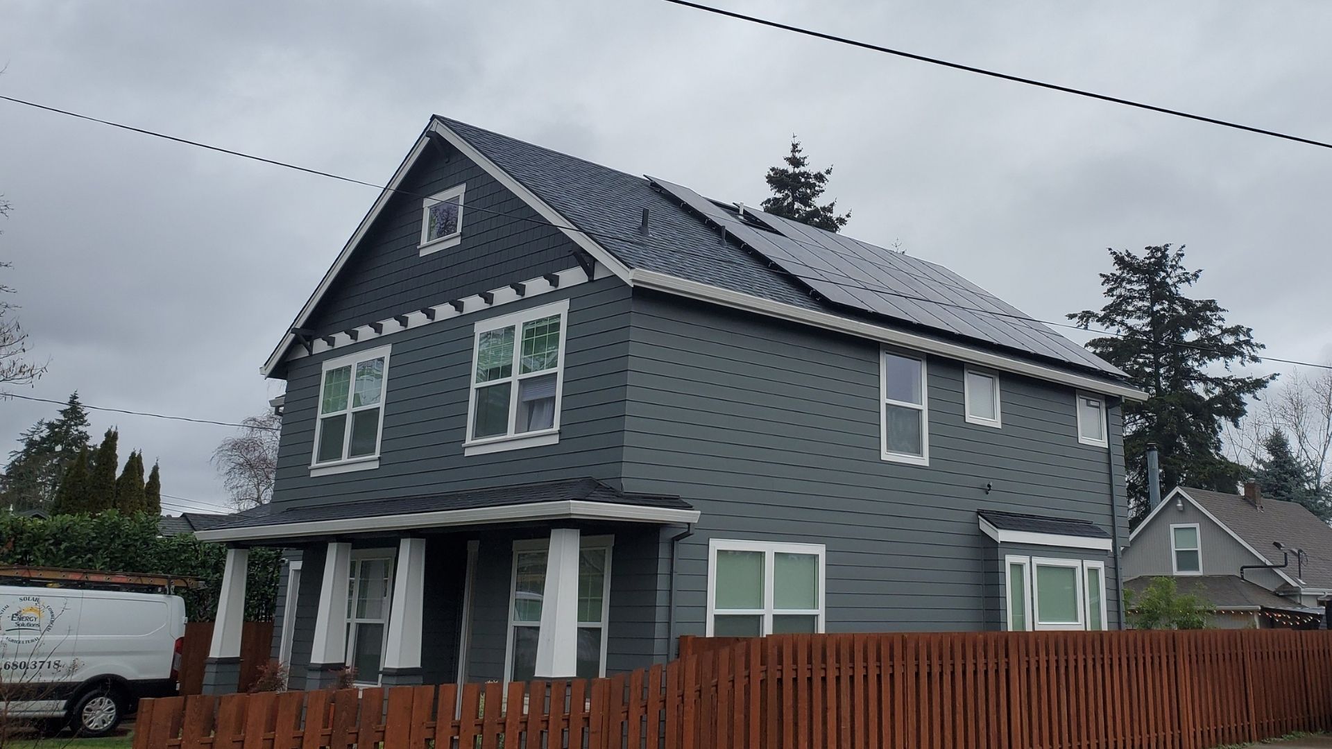 Two-story house with solar panels on roof, gray siding, white trim, and a brown fence.