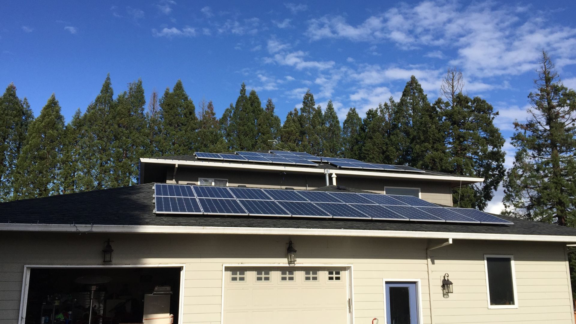 Solar panels installed on a two-story house roof, with trees and blue sky visible.