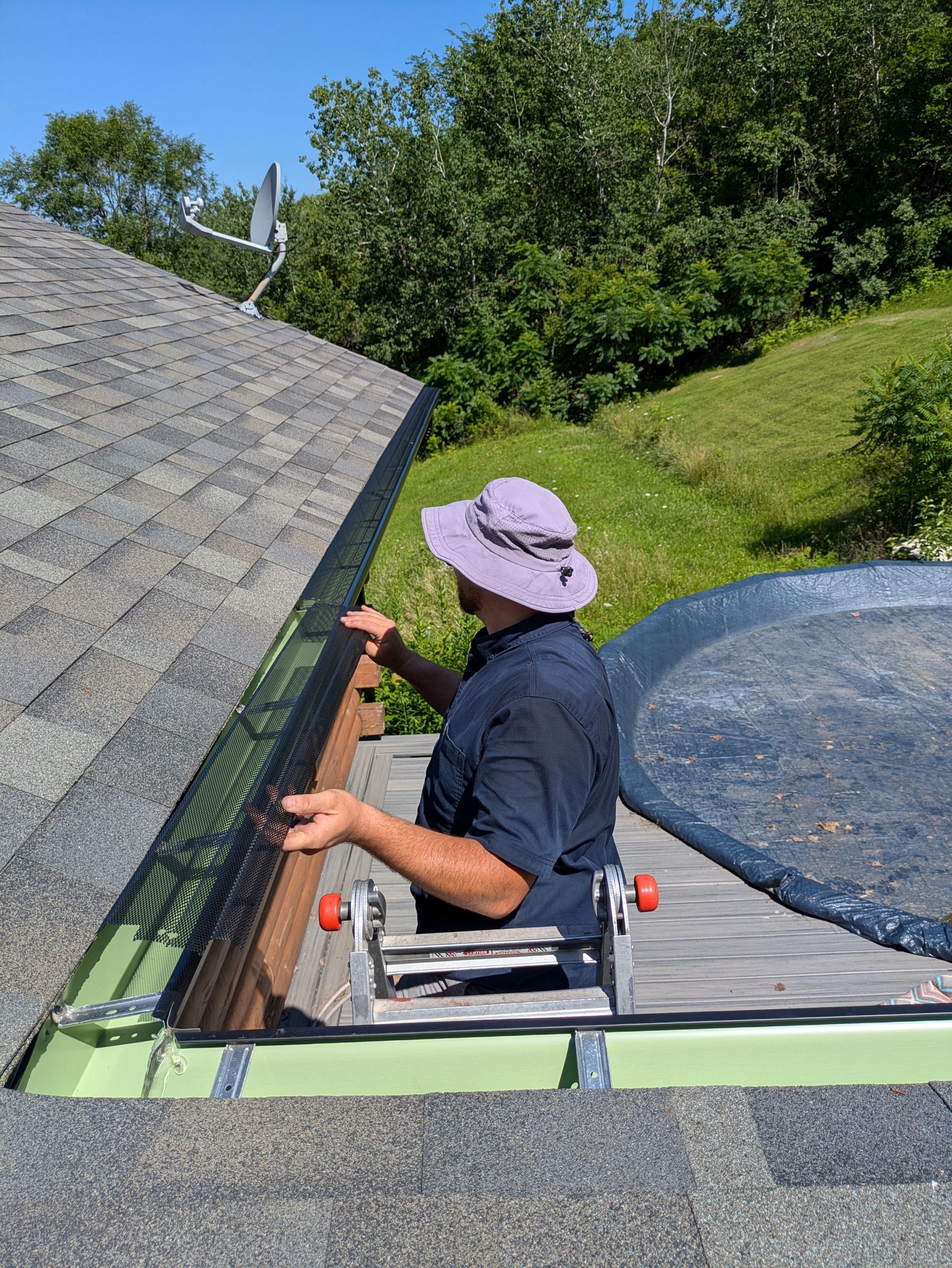 A man is sitting on a ladder cleaning a gutter on a roof.
