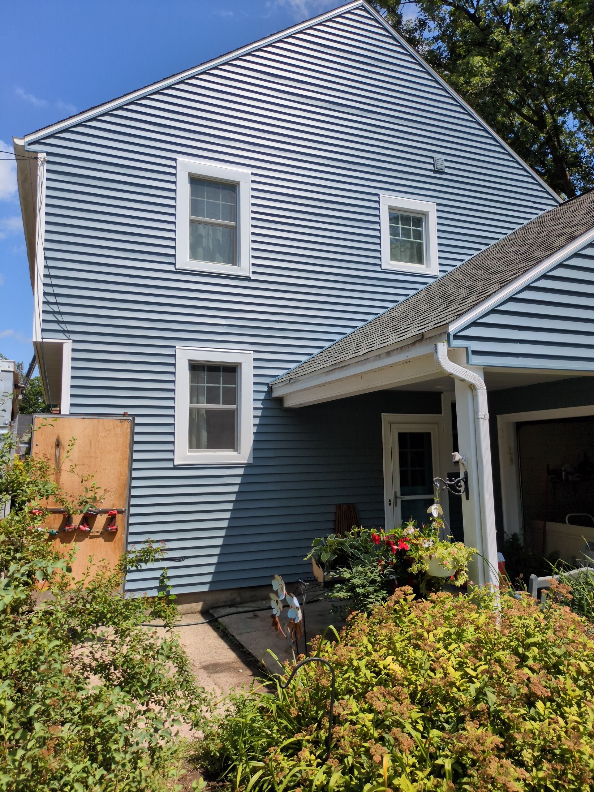 A house with a blue siding and white trim is surrounded by bushes and trees.