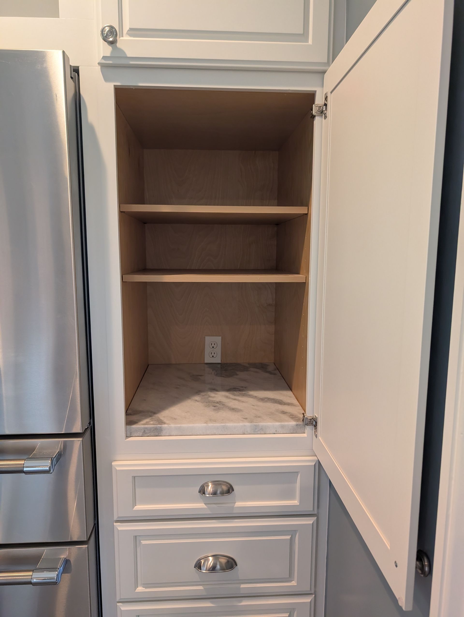 Open white kitchen cabinet with shelves, drawers, and an electrical outlet.