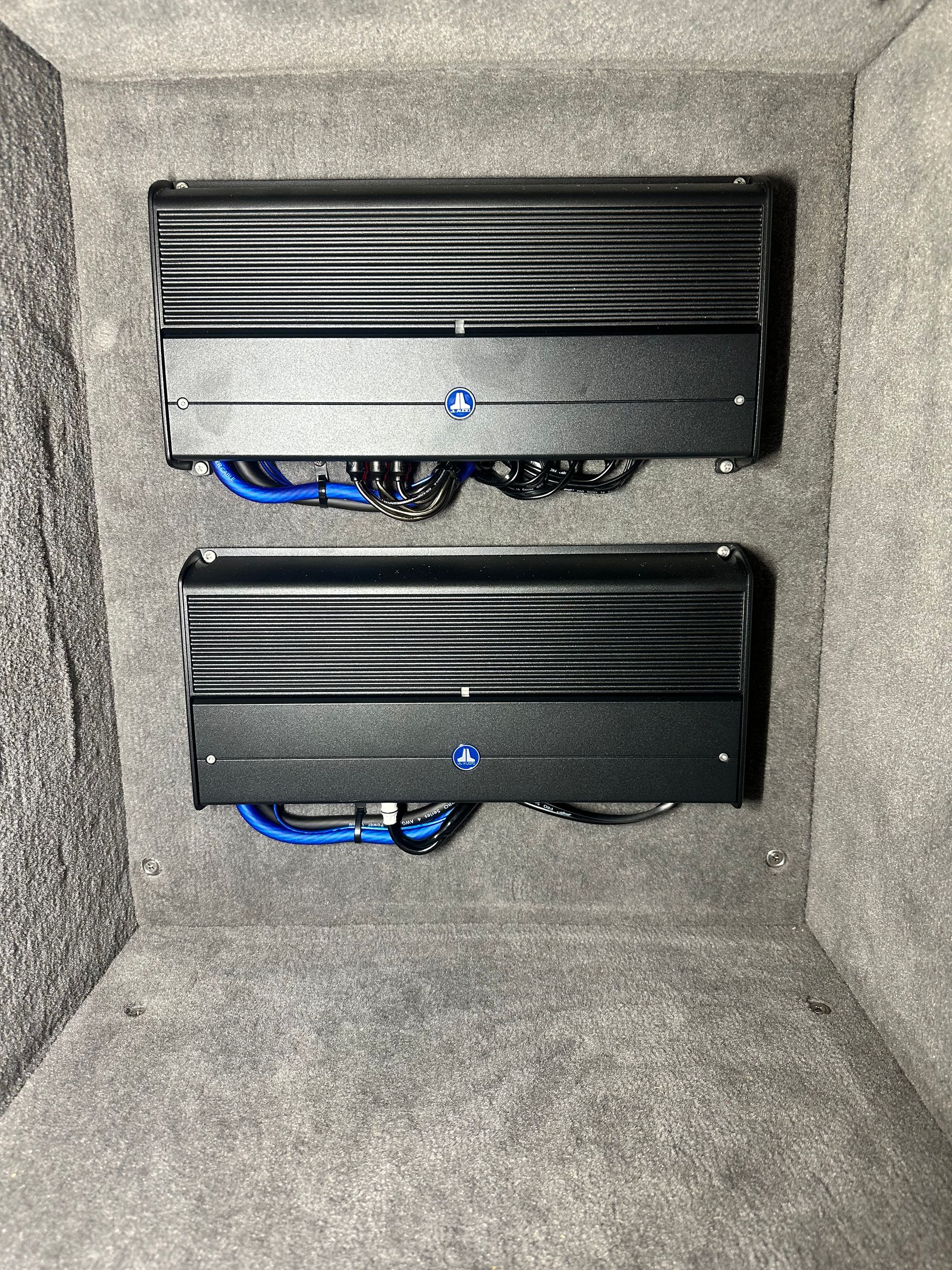 Two black car audio amplifiers mounted in a trunk, with blue wiring visible.