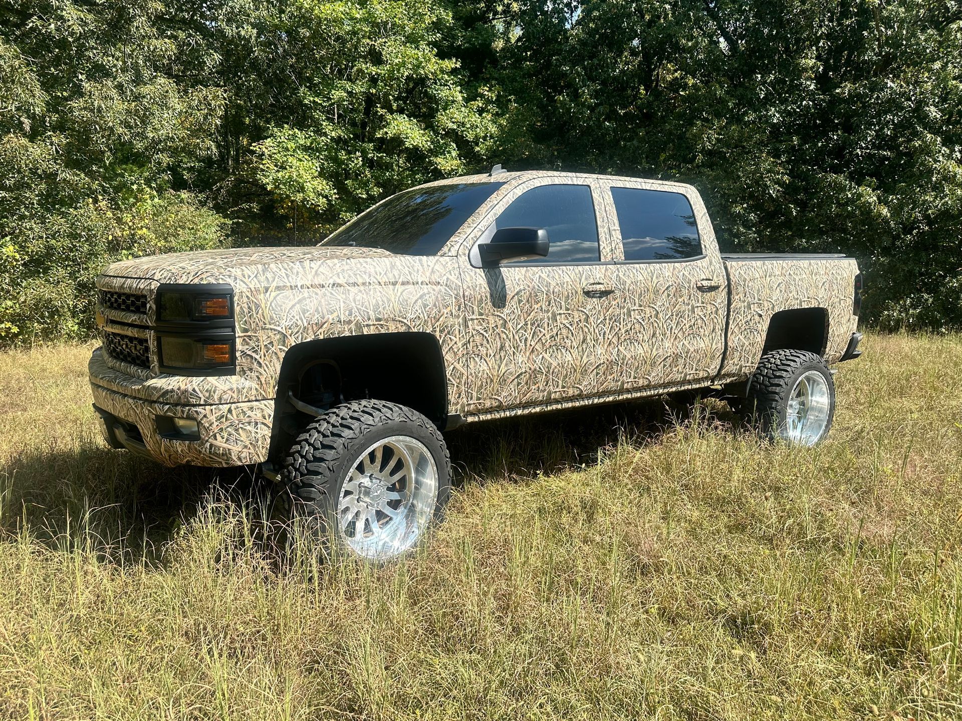 Camouflage pickup truck parked in a grassy field, trees in the background.