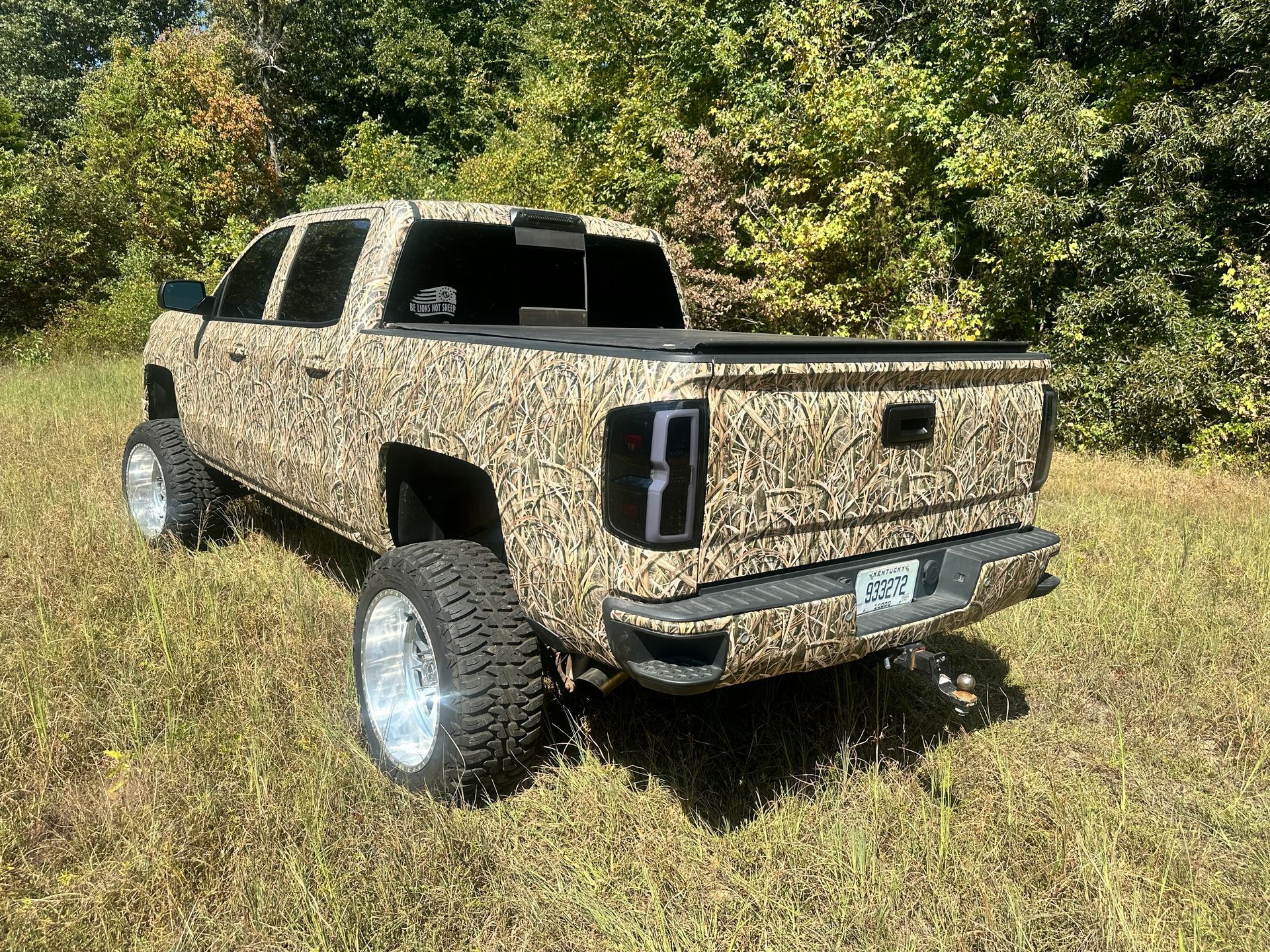Camouflage-wrapped truck with large chrome wheels parked in a grassy field, near trees.