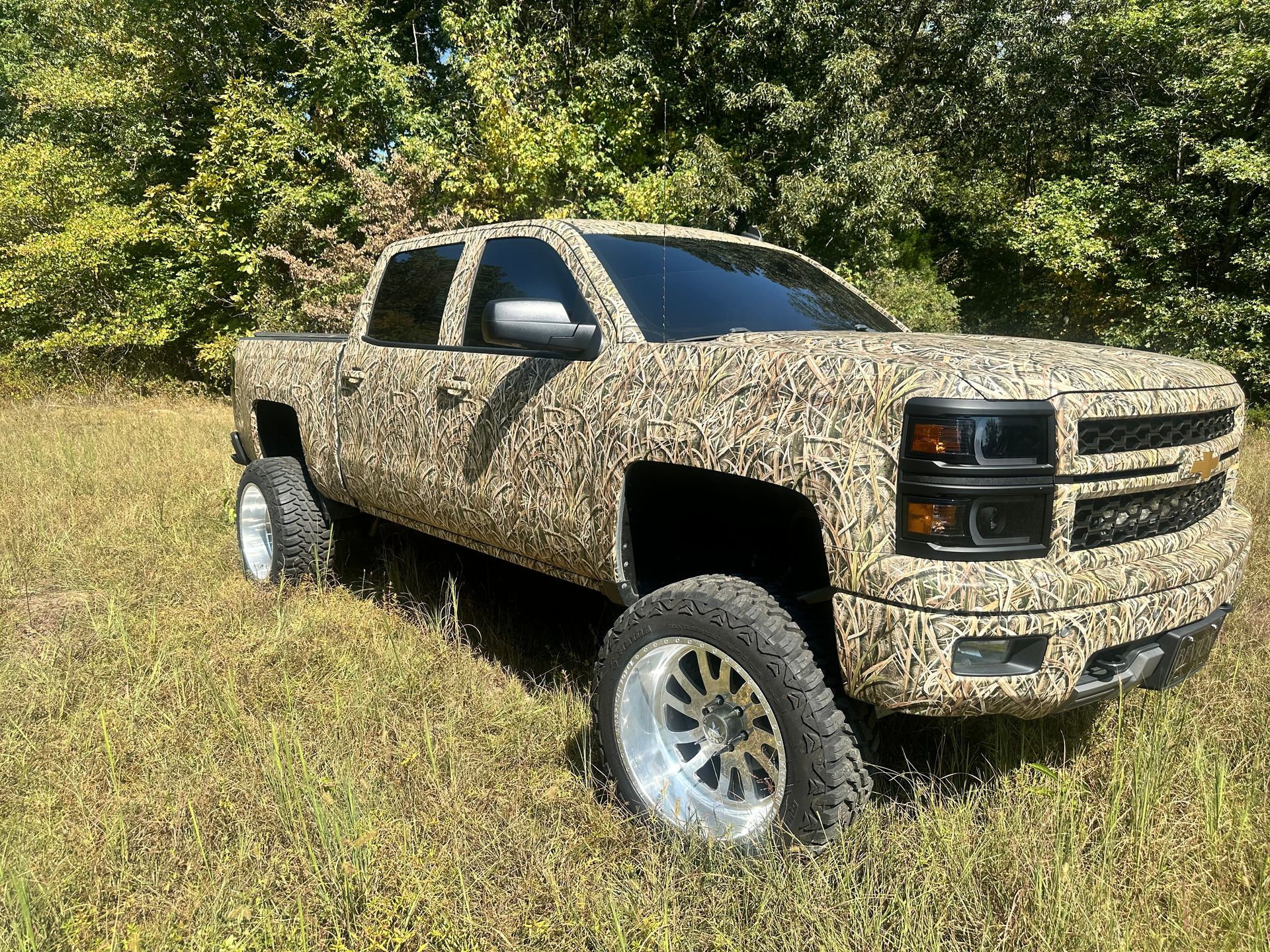 Camouflage-wrapped Chevy Silverado truck on grass, tinted windows, chrome wheels, surrounded by trees.