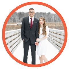 A man in a suit and tie and a woman in a white dress are standing on a bridge holding hands.