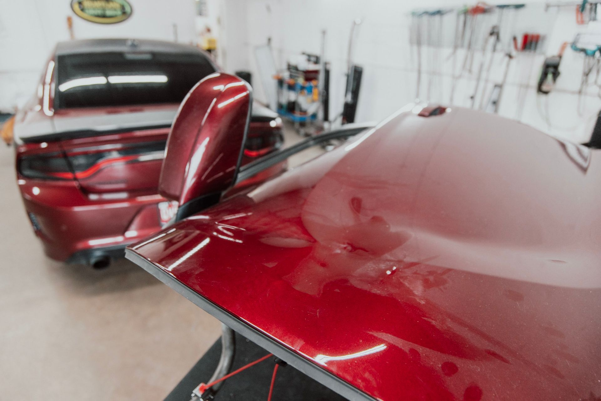 Close-up of a shiny red car's body and side mirror, with another red car in the background in a garage setting.