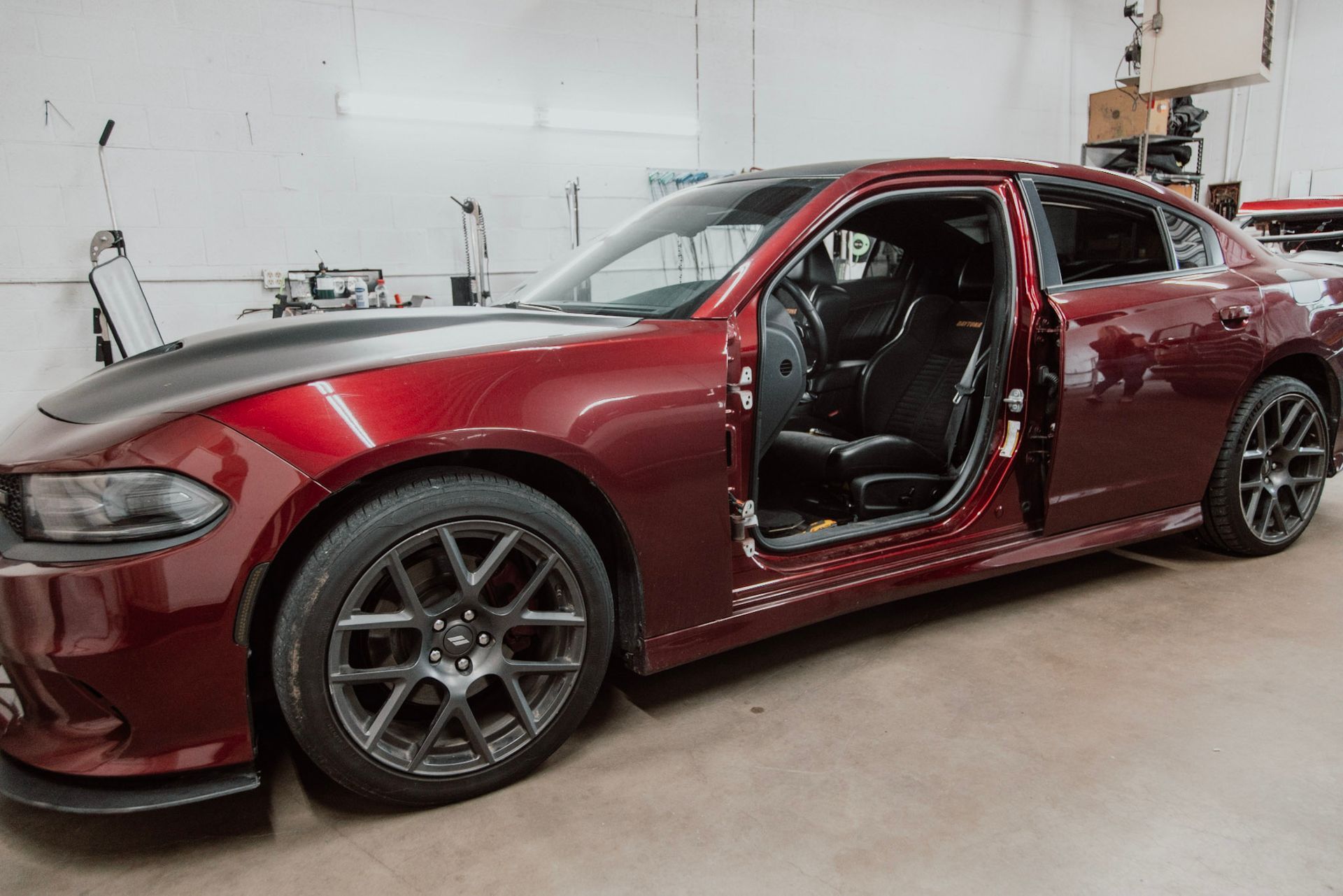 Red Dodge Charger with open doors in a workshop, black wheels and hood, gray walls.