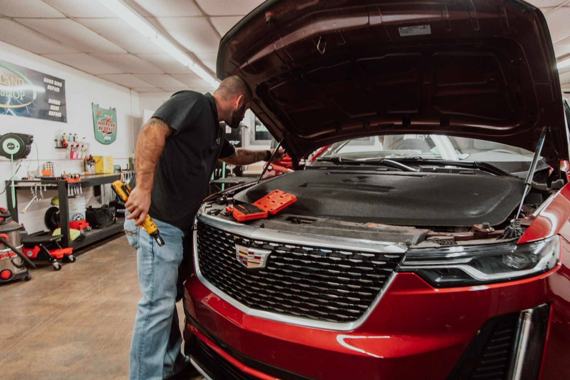 Mechanic working on a red car with the hood open in a garage, using a flashlight and tools.