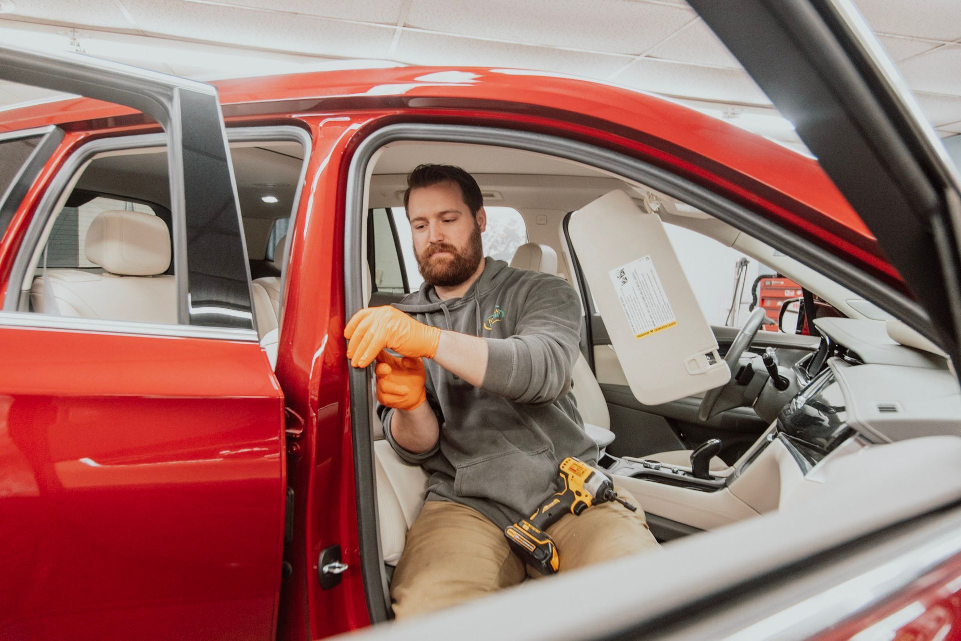 Man in orange gloves works on a red car interior, holding a tool; garage setting.