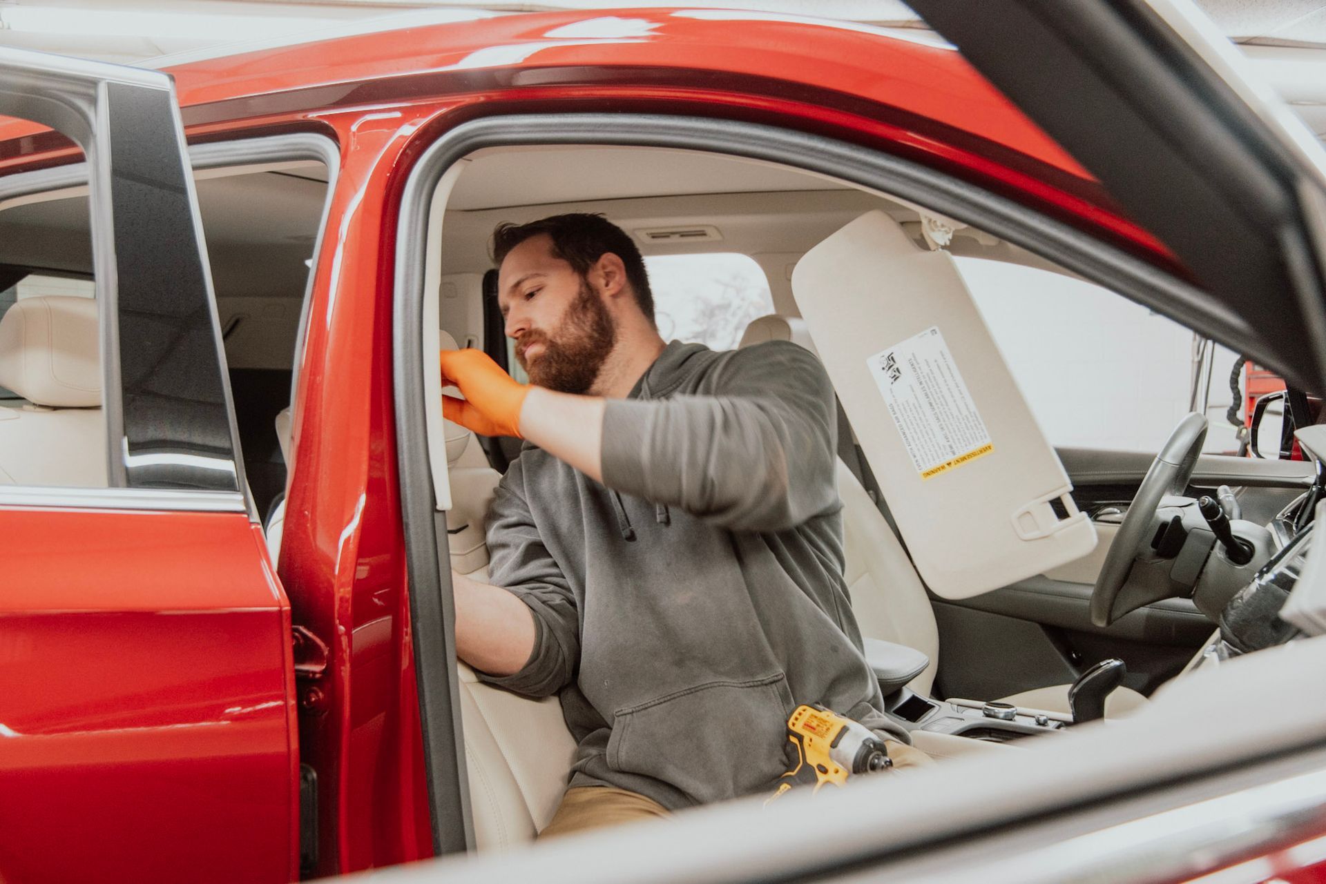 Mechanic working on car's interior; orange gloves, red car, open door, cream seats, visor hanging down.