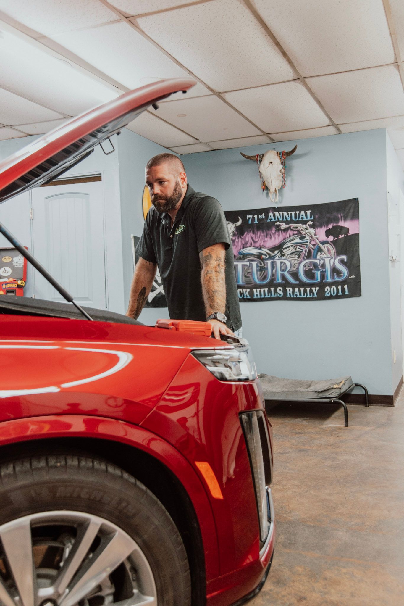 Man in black shirt looking at red car's open hood in a garage, Sturgis banner in background.