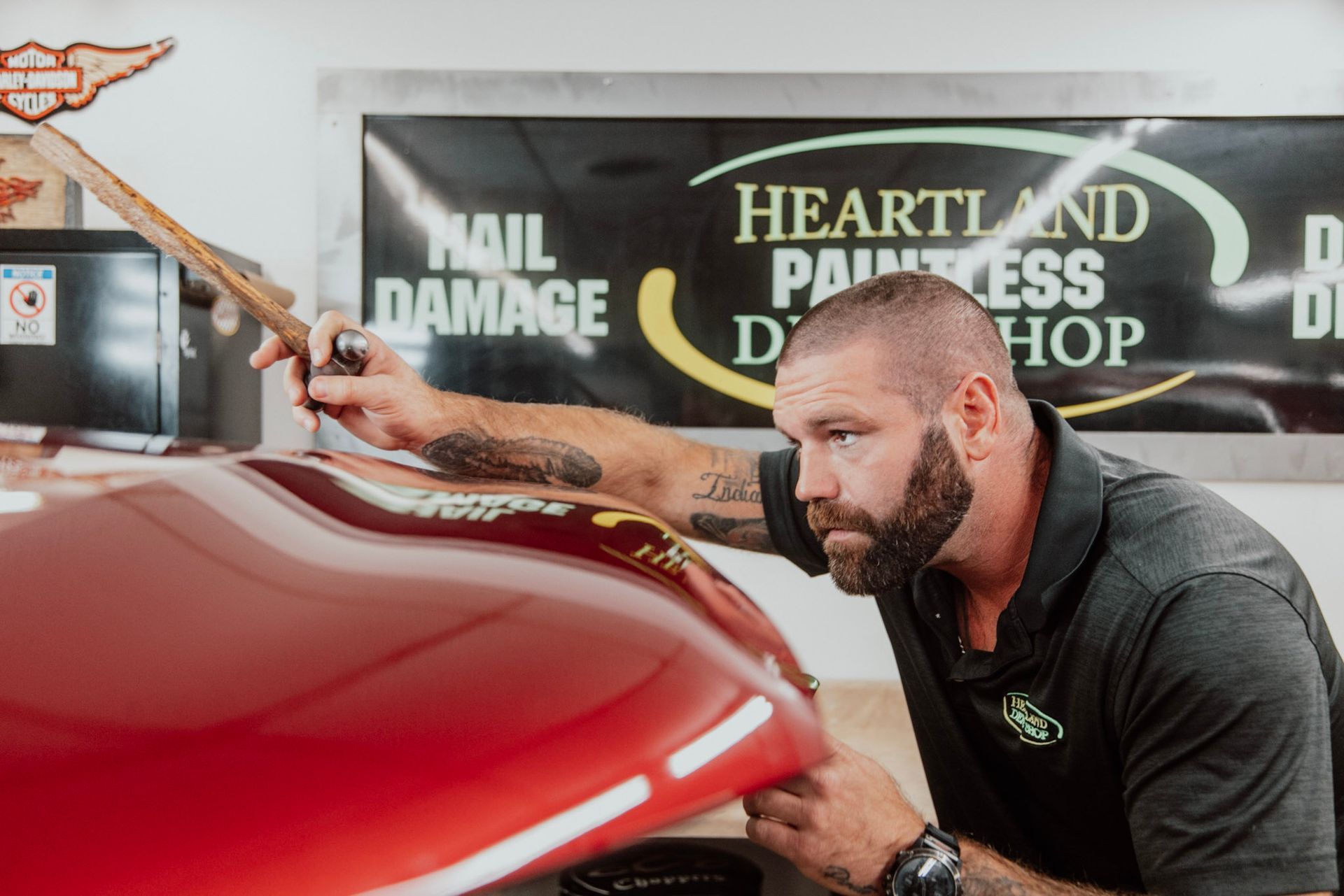 Man examines red car surface in a Heartland Paintless Dent Repair shop, holding a tool, focused.