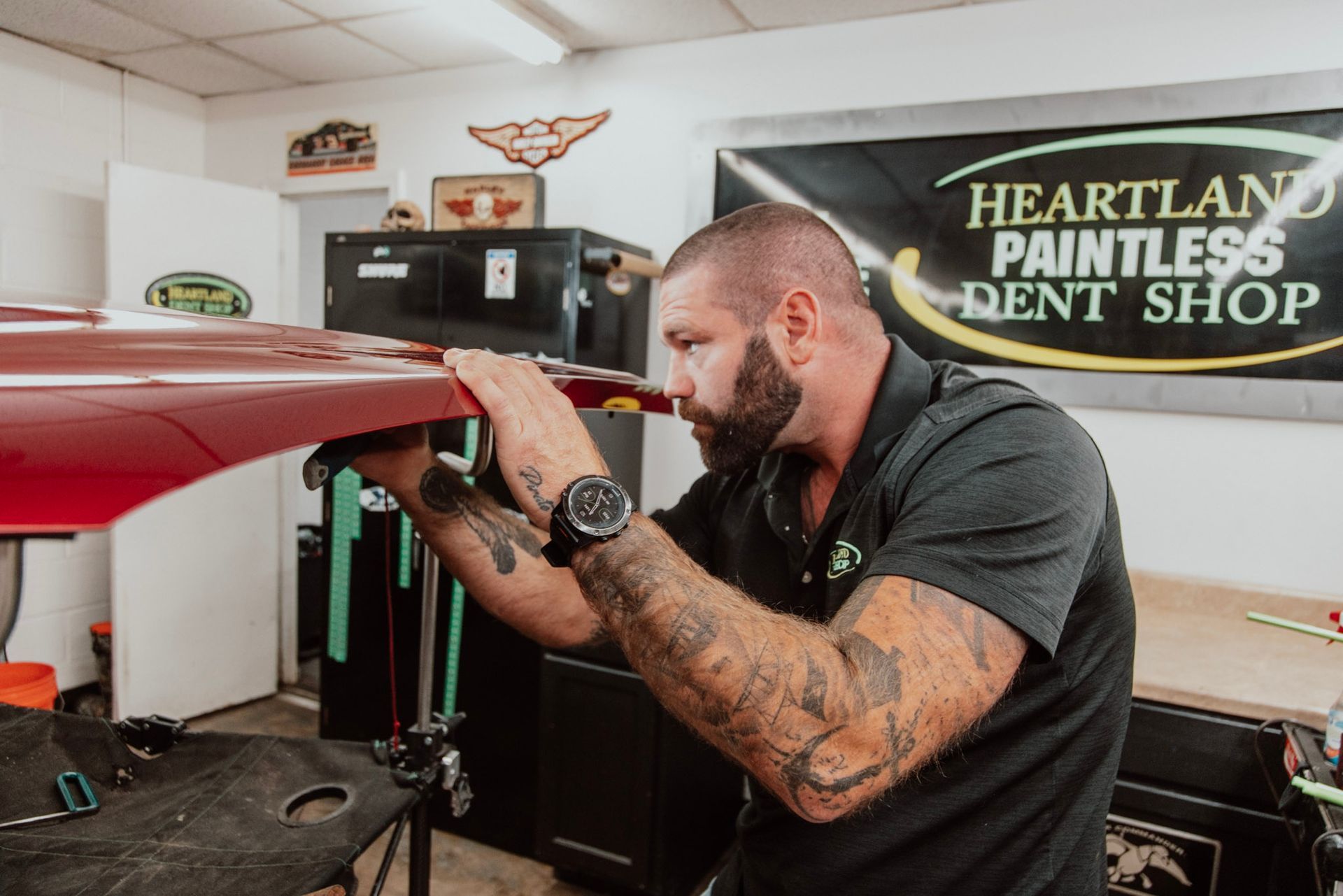 Man inspecting a red car panel at Heartland Paintless Dent Shop. He is in a garage, looking focused.