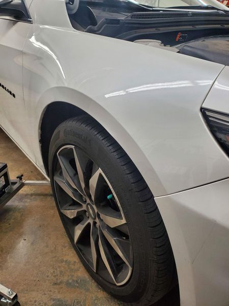 White car's front side and tire detail, with a dark wheel. The setting appears to be indoors.