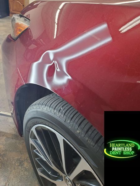 Red car with dented fender, illuminated by reflection, near tire. Heartland Paintless Dent Shop logo.